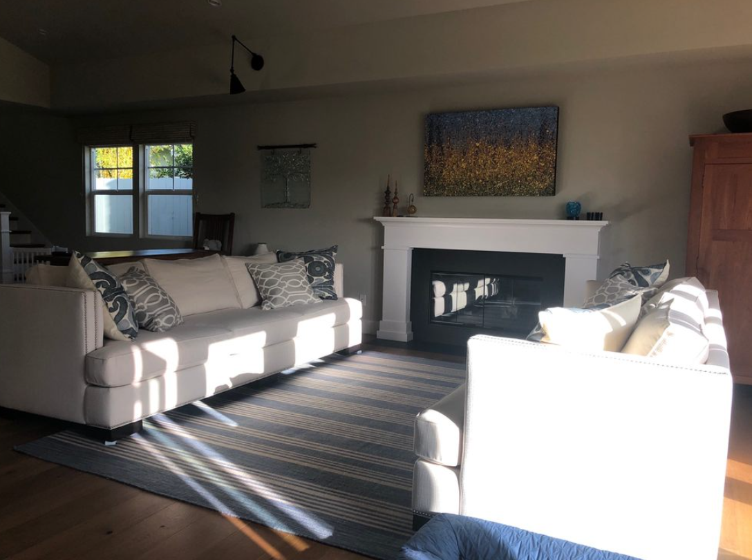 Sunlit living room with white sofas, patterned rug, fireplace, and wooden cabinet