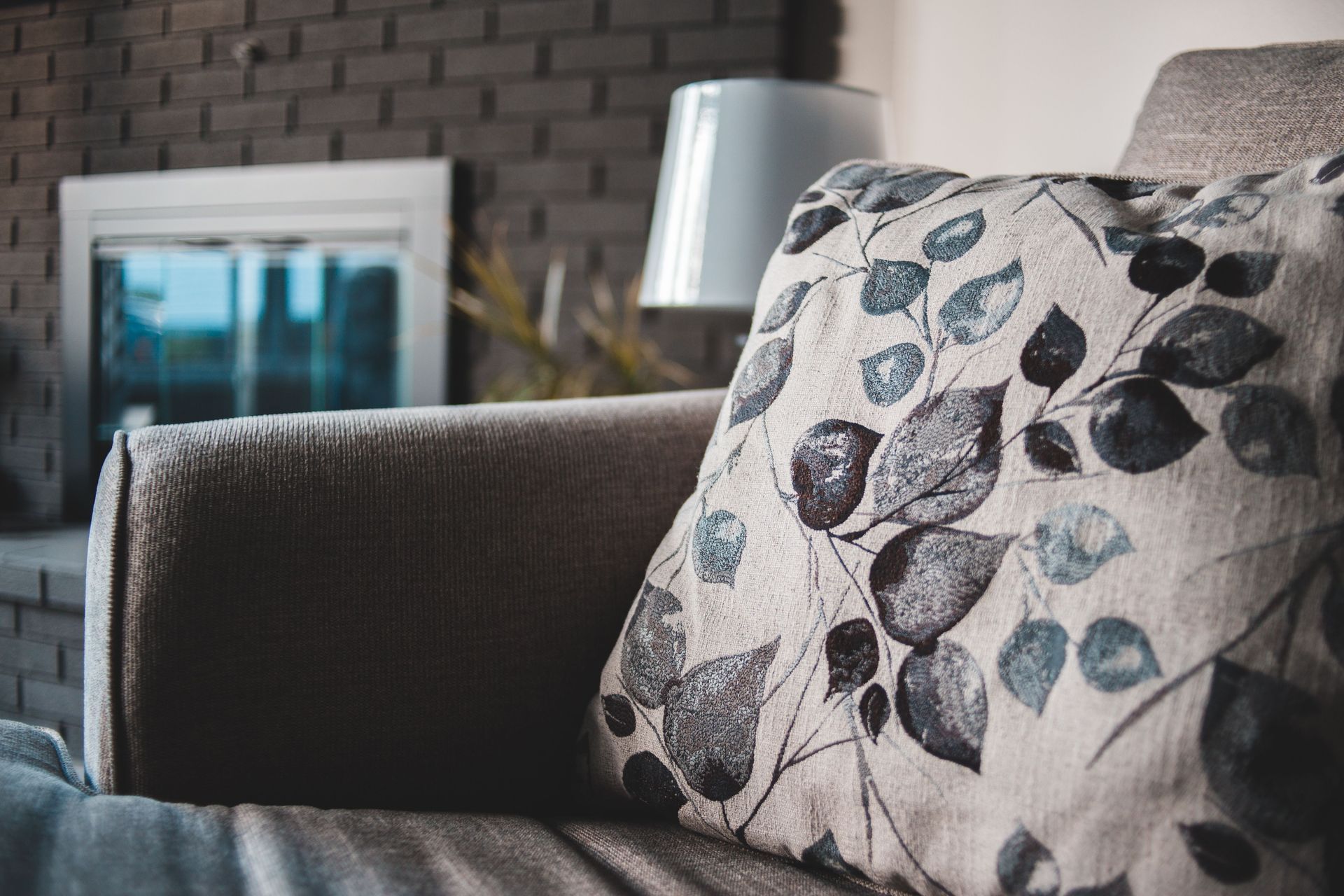 Gray sofa with a floral pillow beside a white lamp against a dark brick wall