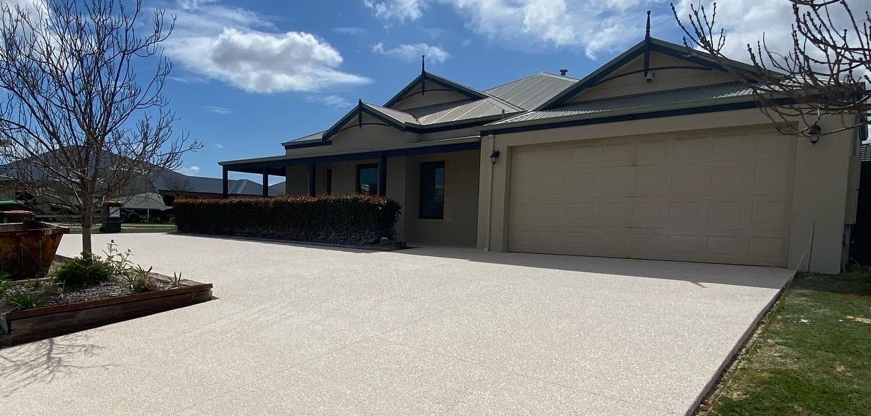 Light coloured exposed aggregate driveway leading to and wrapping around modern home with landscaping.
