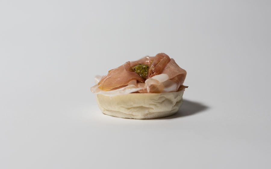 Antipasto: base di pasta ricoperta di prosciutto e pesto verde su una superficie bianca.