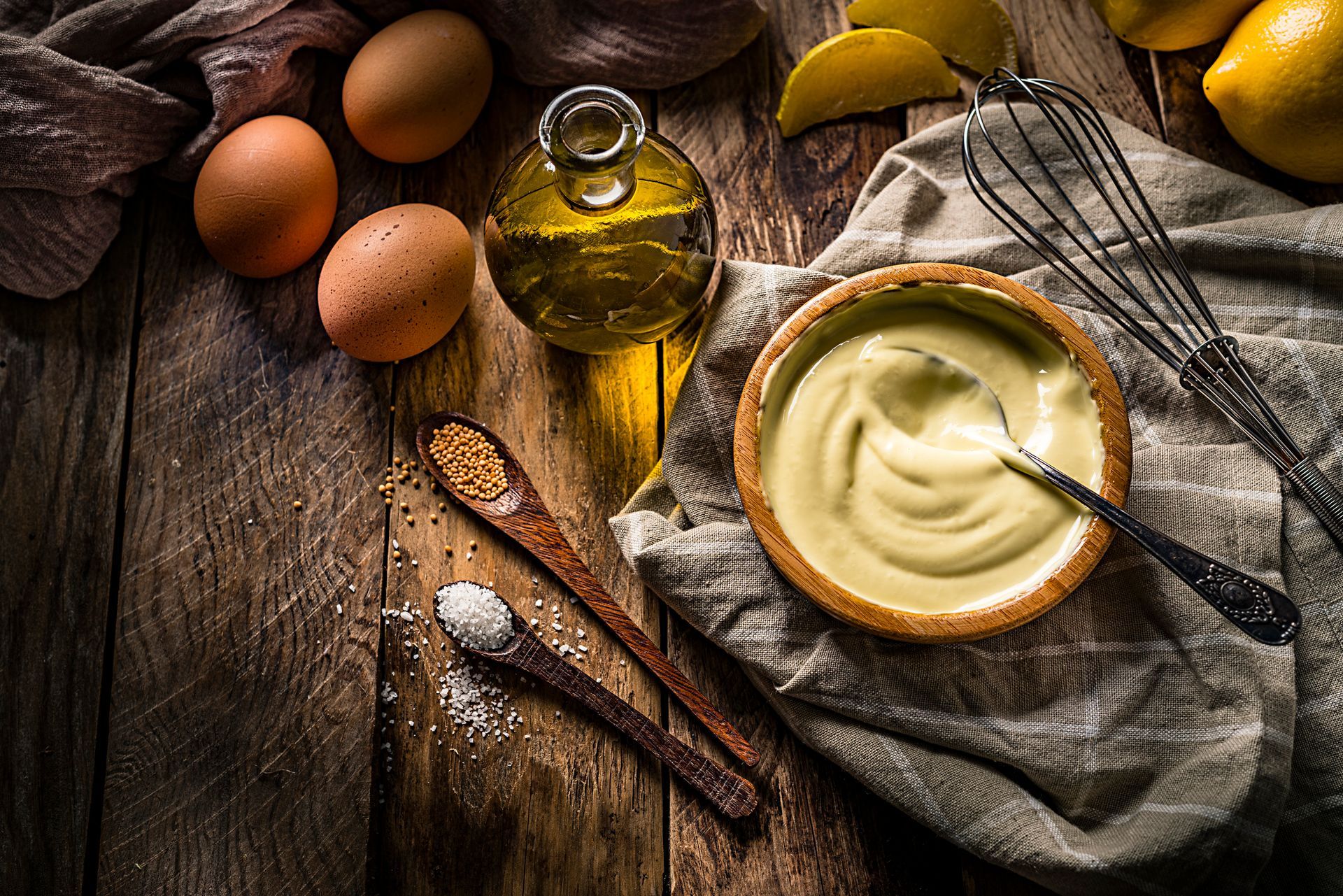Ingredienti della maionese: uova, olio, limone e spezie disposti su un tavolo di legno.