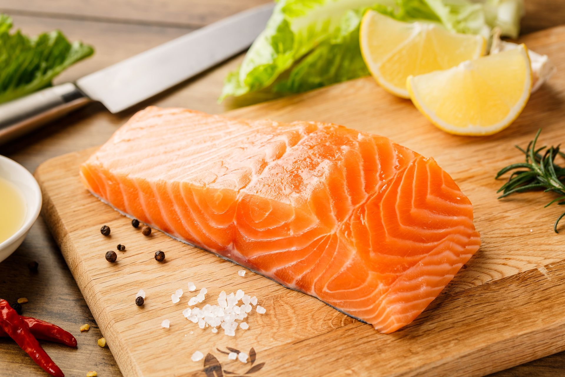 Filetto di salmone crudo su un tagliere di legno con fette di limone, spezie e un coltello.