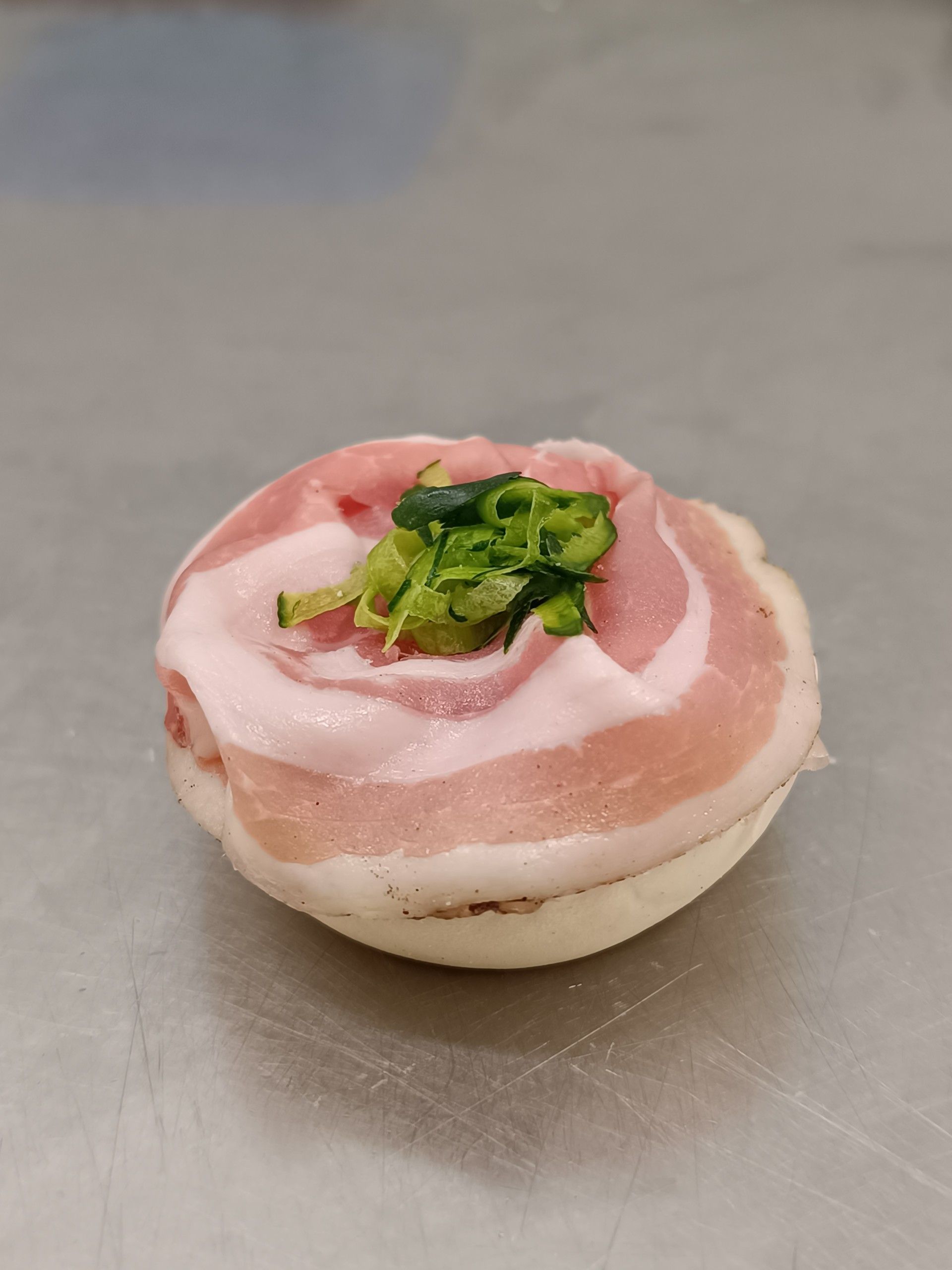 Pancetta di maiale avvolta attorno a un pezzo bianco e rotondo, guarnita con cipollotti verdi tritati, su una superficie grigia.