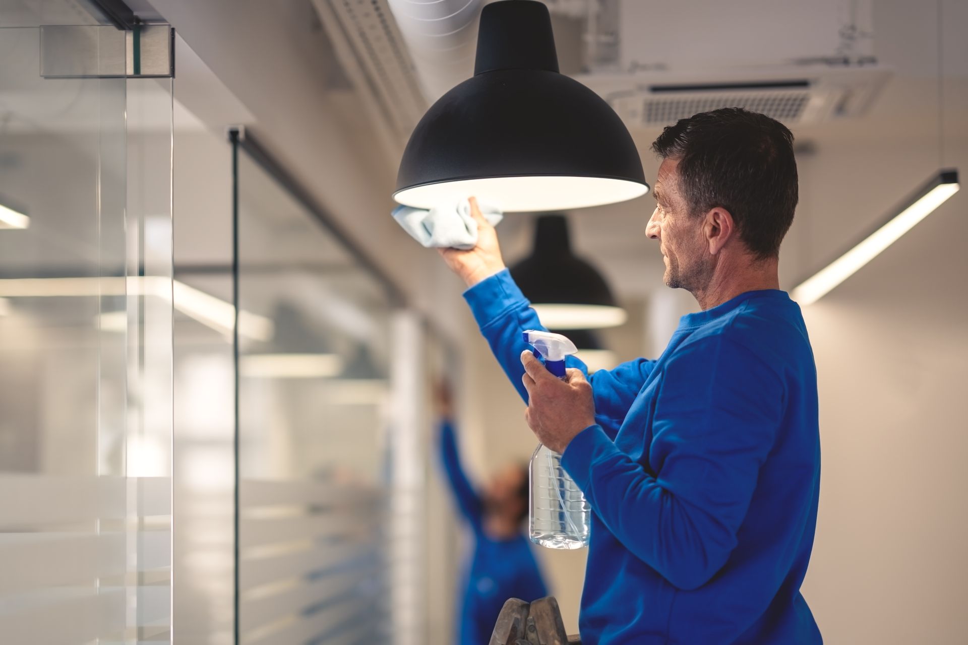 Technician replacing a light bulb under a black pendant lamp in a bright office hallway