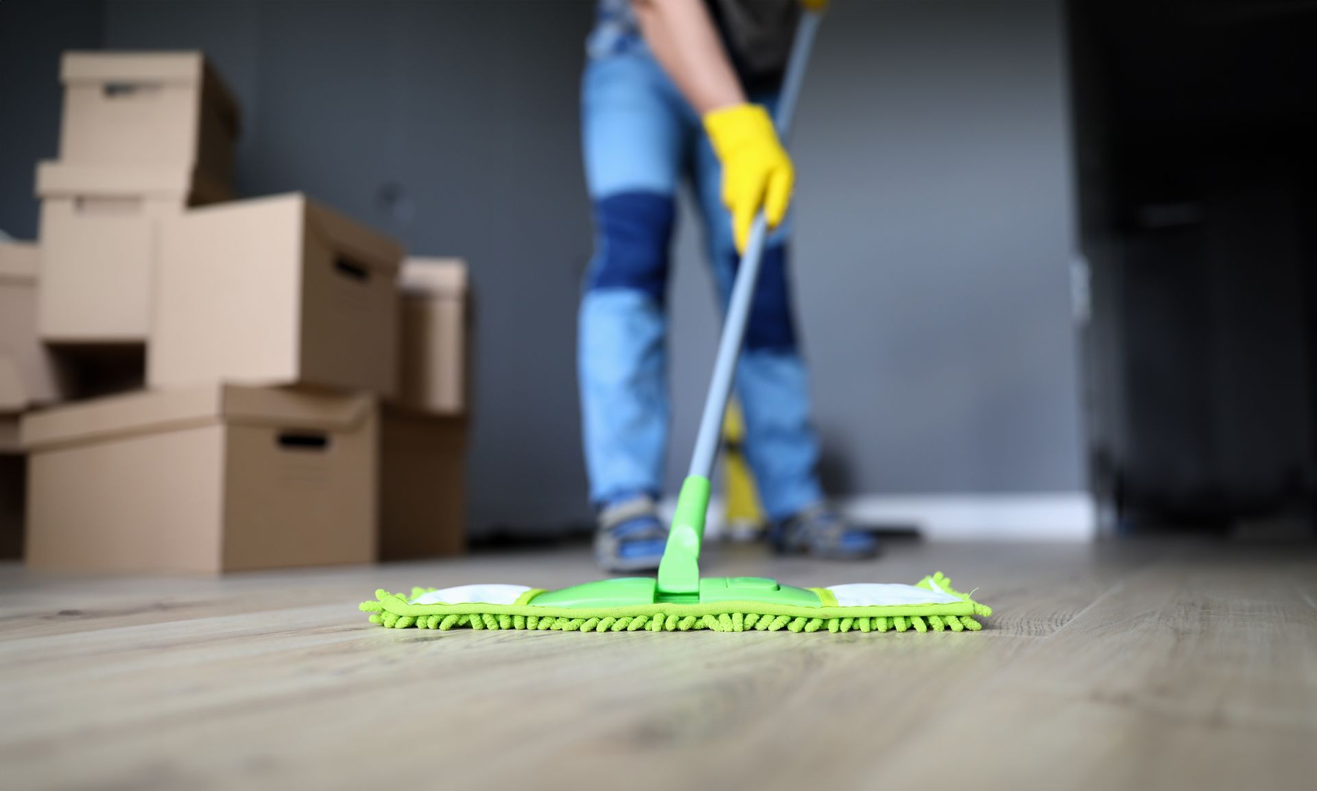 Person mopping a wooden floor with a green mop beside stacked moving boxes