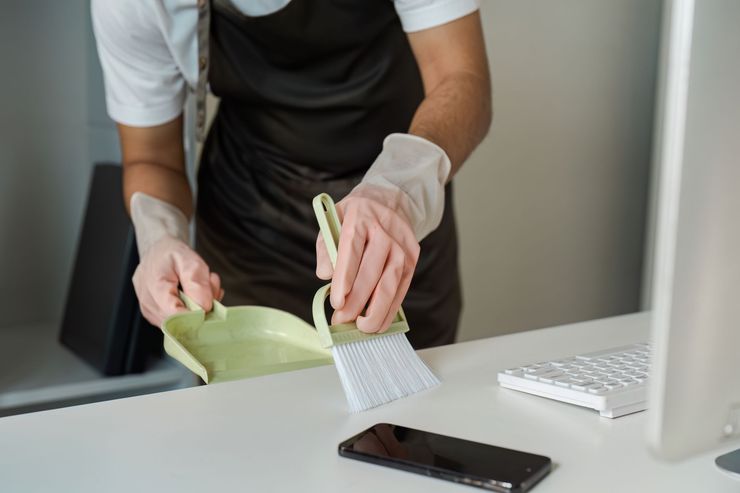 Gloved person cleaning a desk with a dustpan and brush beside a phone and keyboard