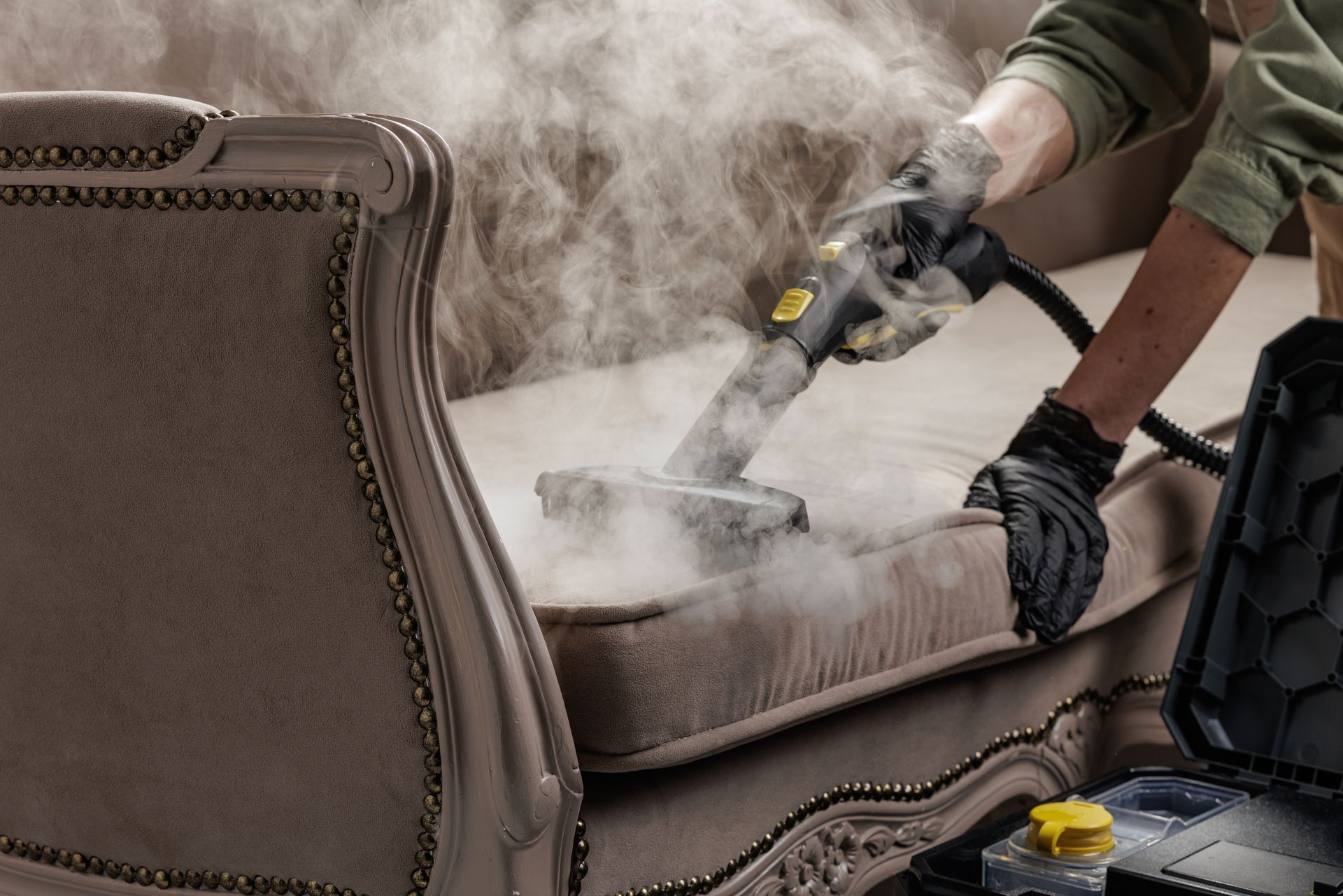 Person steam-cleans a beige sofa, lifting dirt as thick white steam fills the room.