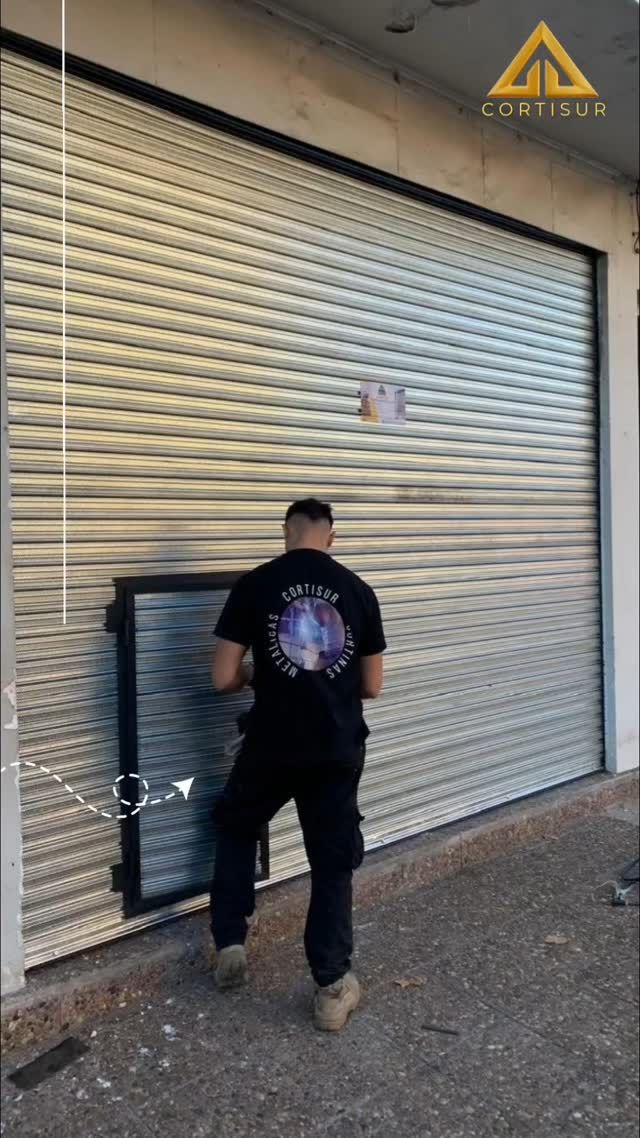Un hombre intenta abrir una persiana metálica de seguridad. Viste camisa y pantalones negros, afuera de un edificio.