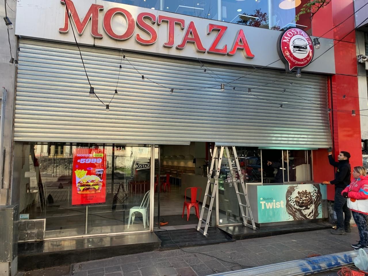 Exterior del restaurante 'Mostaza' con persianas cerradas, un cartel rojo y trabajadores. Dentro hay mesas y sillas.