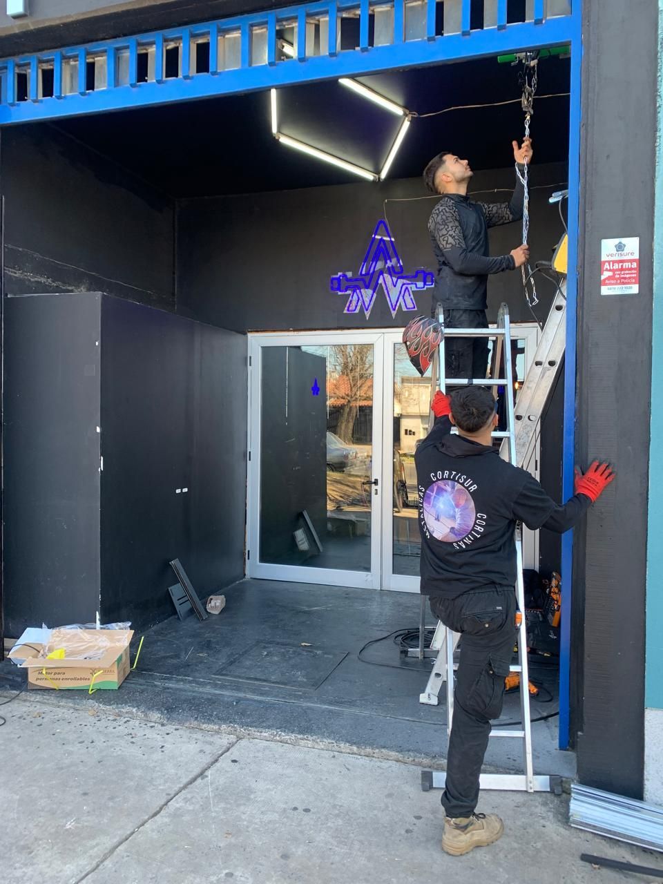 Dos hombres instalando accesorios para escaparates, uno en una escalera, fachada negra y azul.