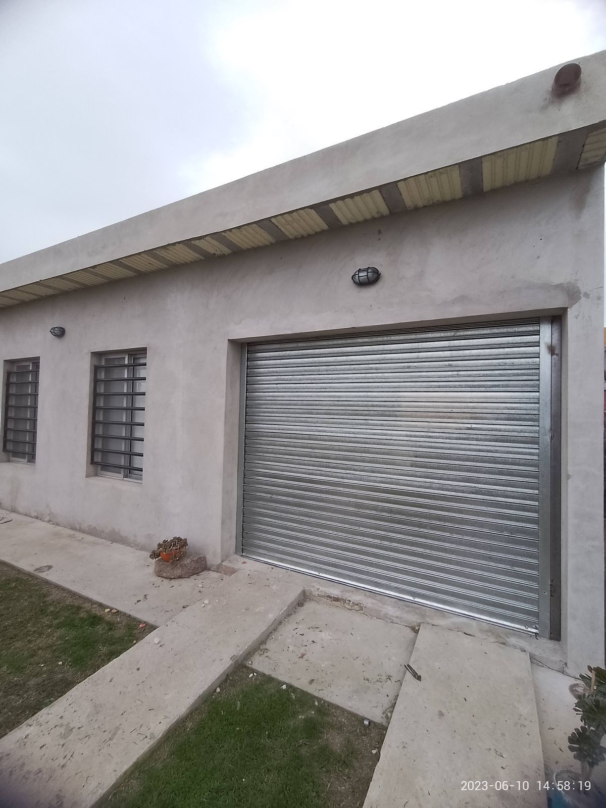 Edificio gris con puerta metálica enrollable, dos ventanas enrejadas y camino de hormigón que conduce a él.