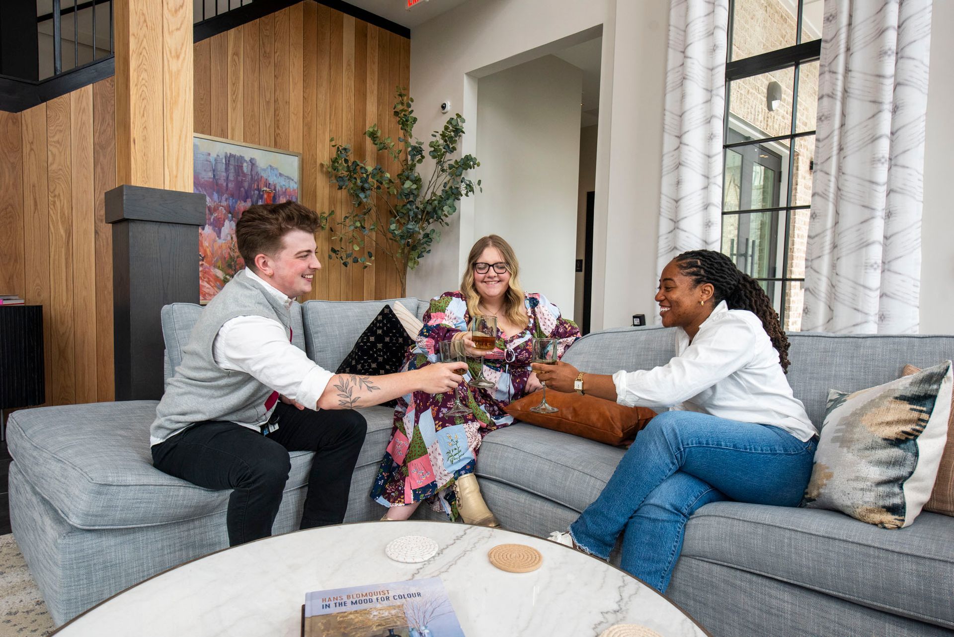 Three people toasting drinks on a gray sectional sofa in a living room.