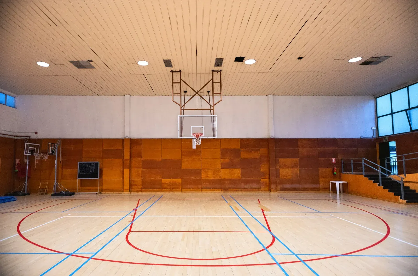 An empty indoor basketball court featuring a wood-paneled wall, light-colored wooden floor, and a single hoop.