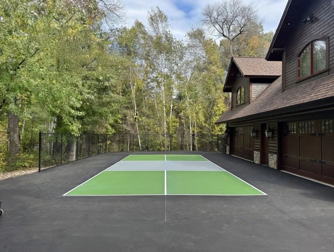 Outdoor pickleball court beside a wooden house, with green and gray playing surface surrounded by trees