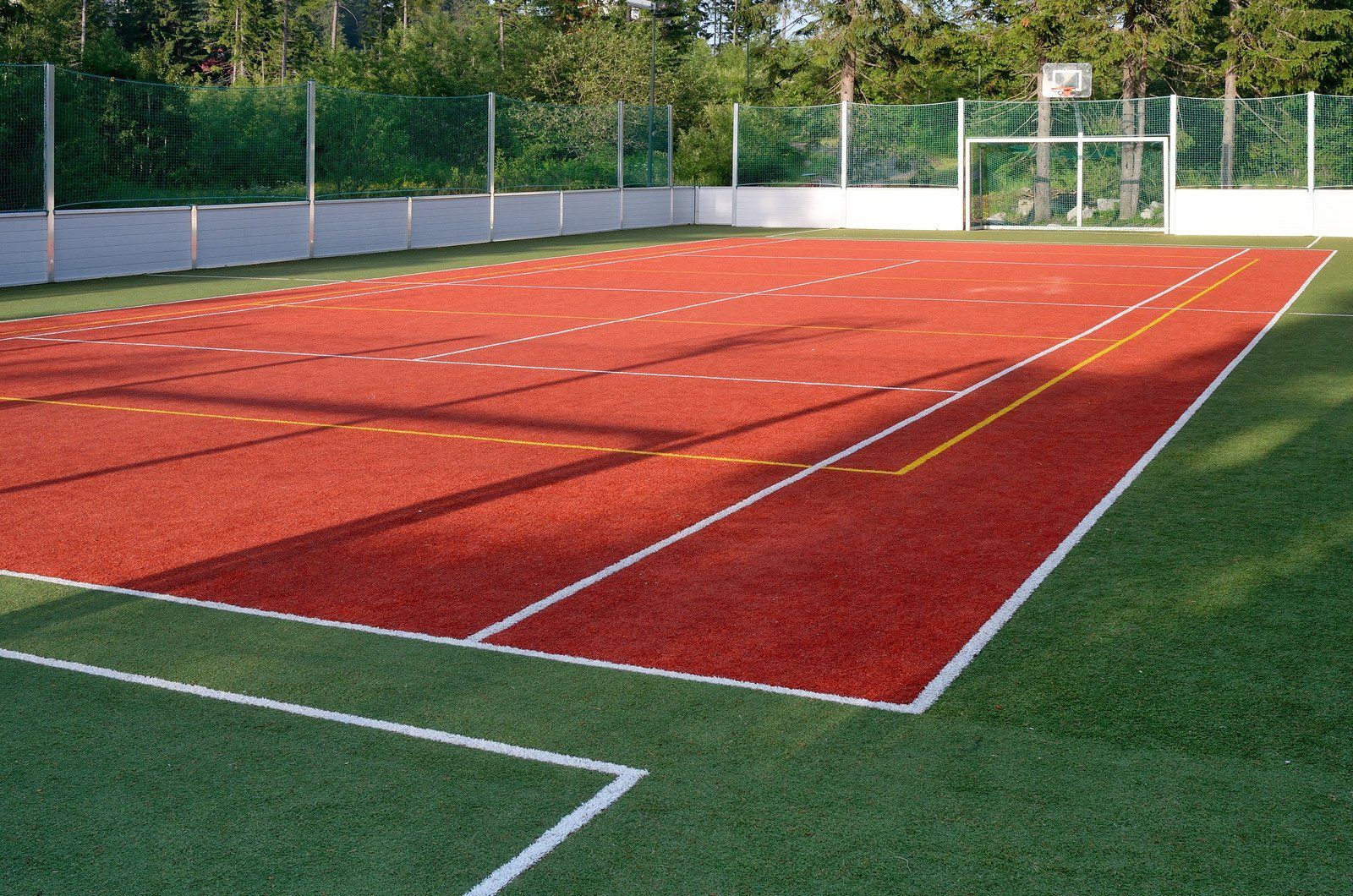 Red sports court with white lines and green perimeter, basketball hoop, soccer goal.