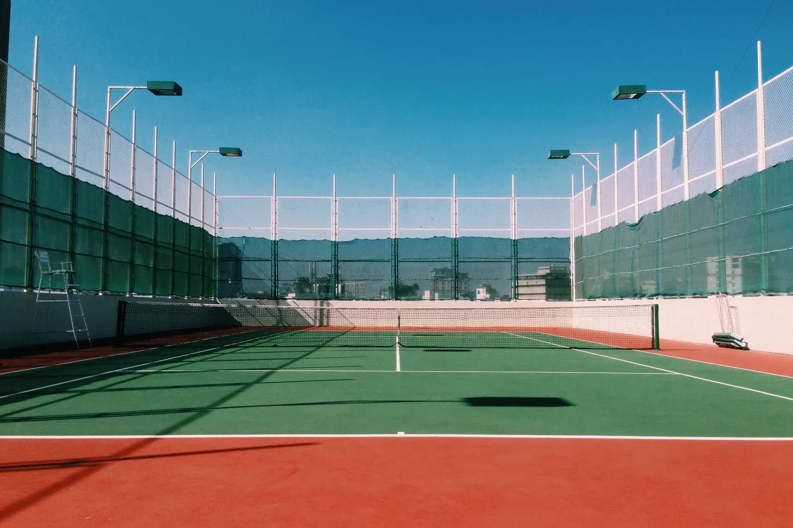 Tennis court with green and red surface, enclosed by fencing, under a blue sky.