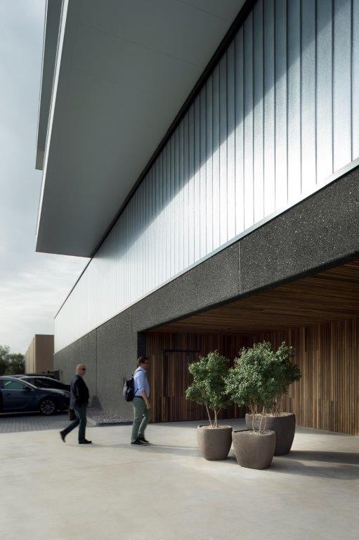Modern building entrance with two people walking, potted trees, and a parked car nearby