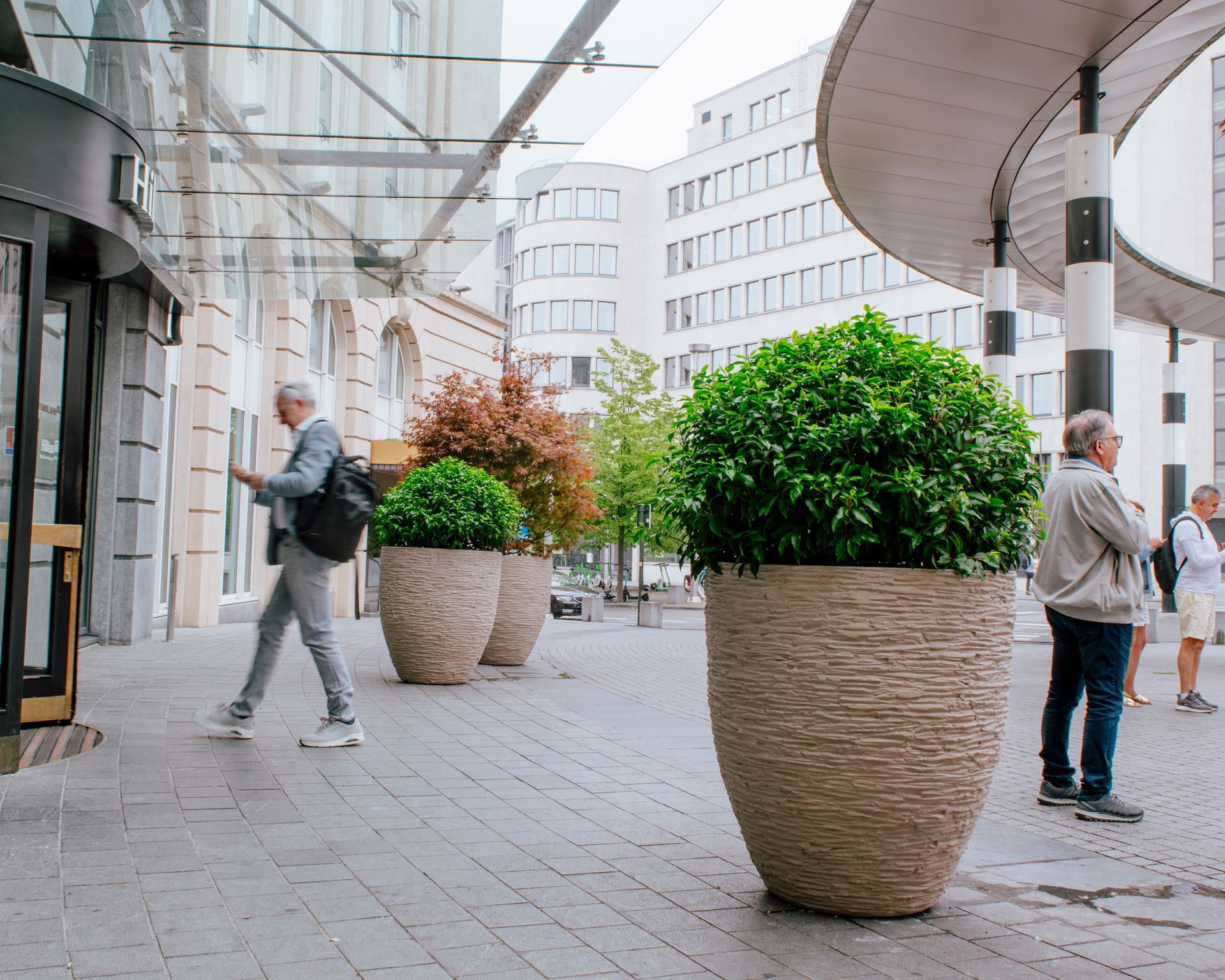 Modern shopping street with large potted shrubs and a few pedestrians under a glass canopy