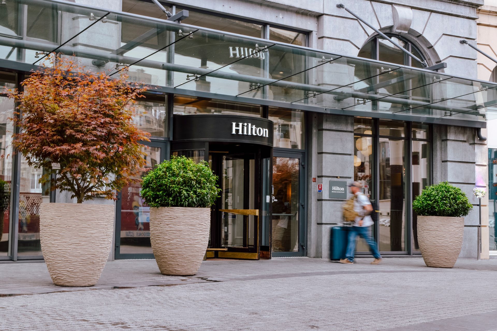 Hilton hotel entrance with glass canopy, potted trees, and a person standing by the door
