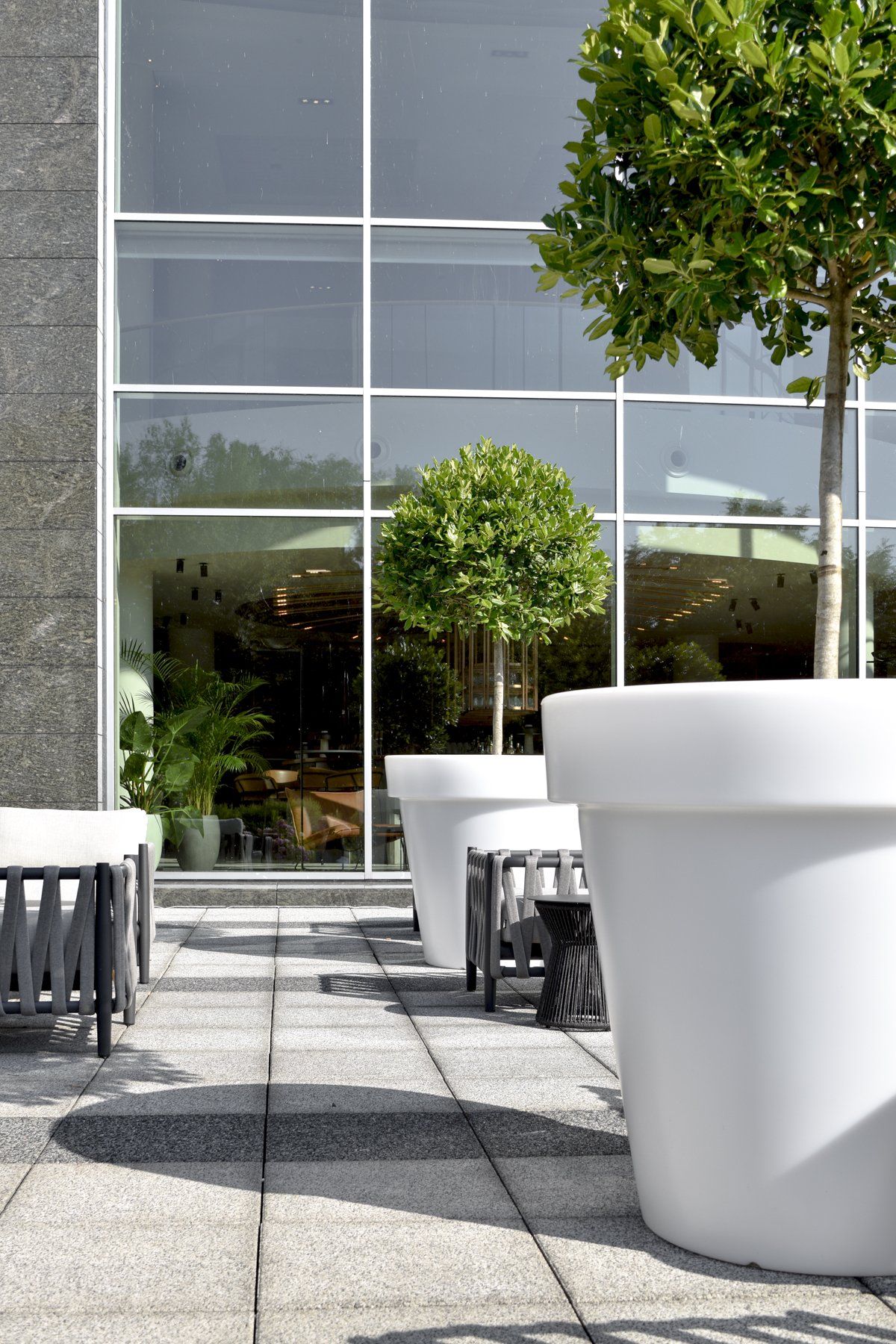 Outdoor patio with white tables, chairs, and large planters beside a glass building.