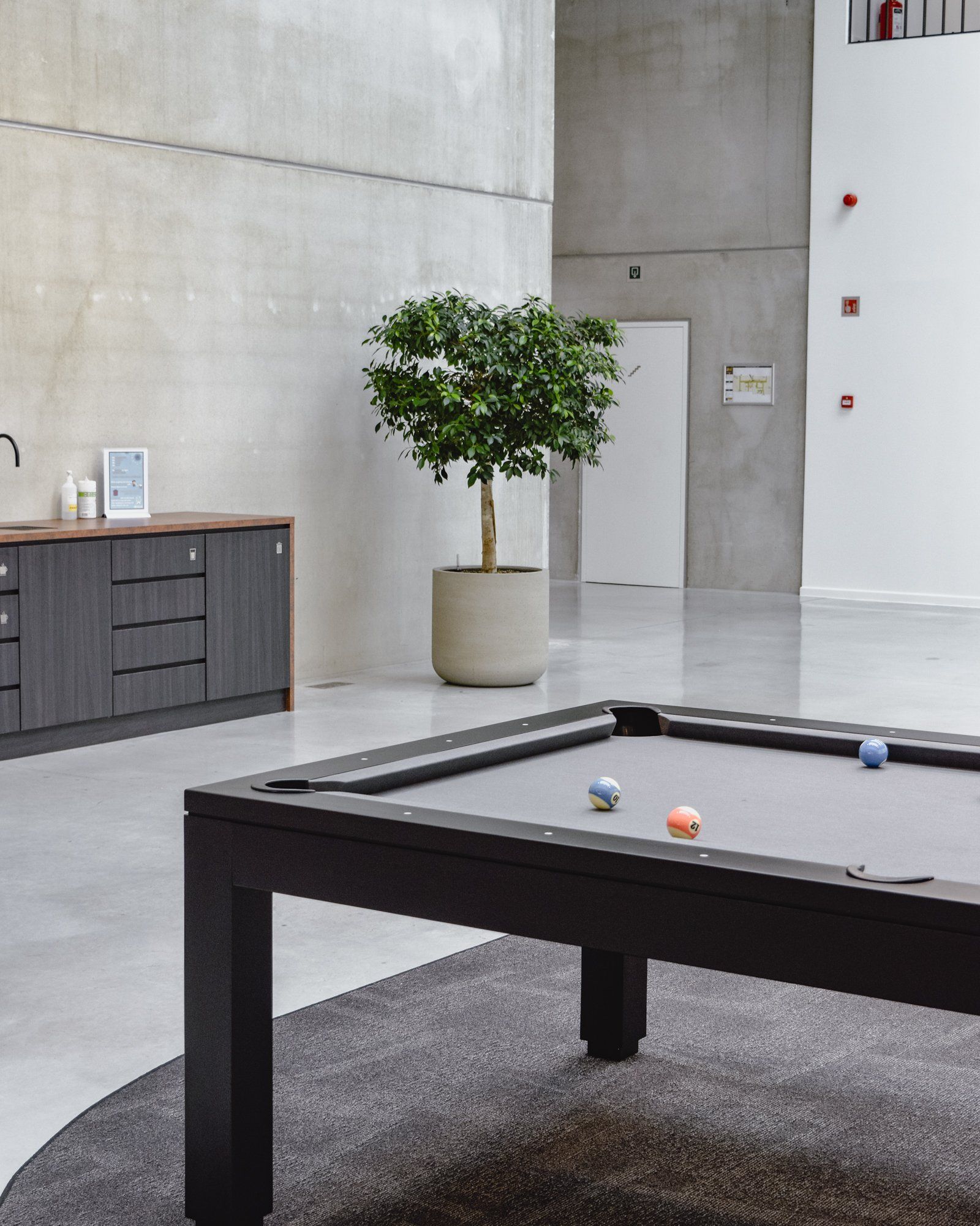Modern lounge with a pool table, potted tree, and gray concrete walls.