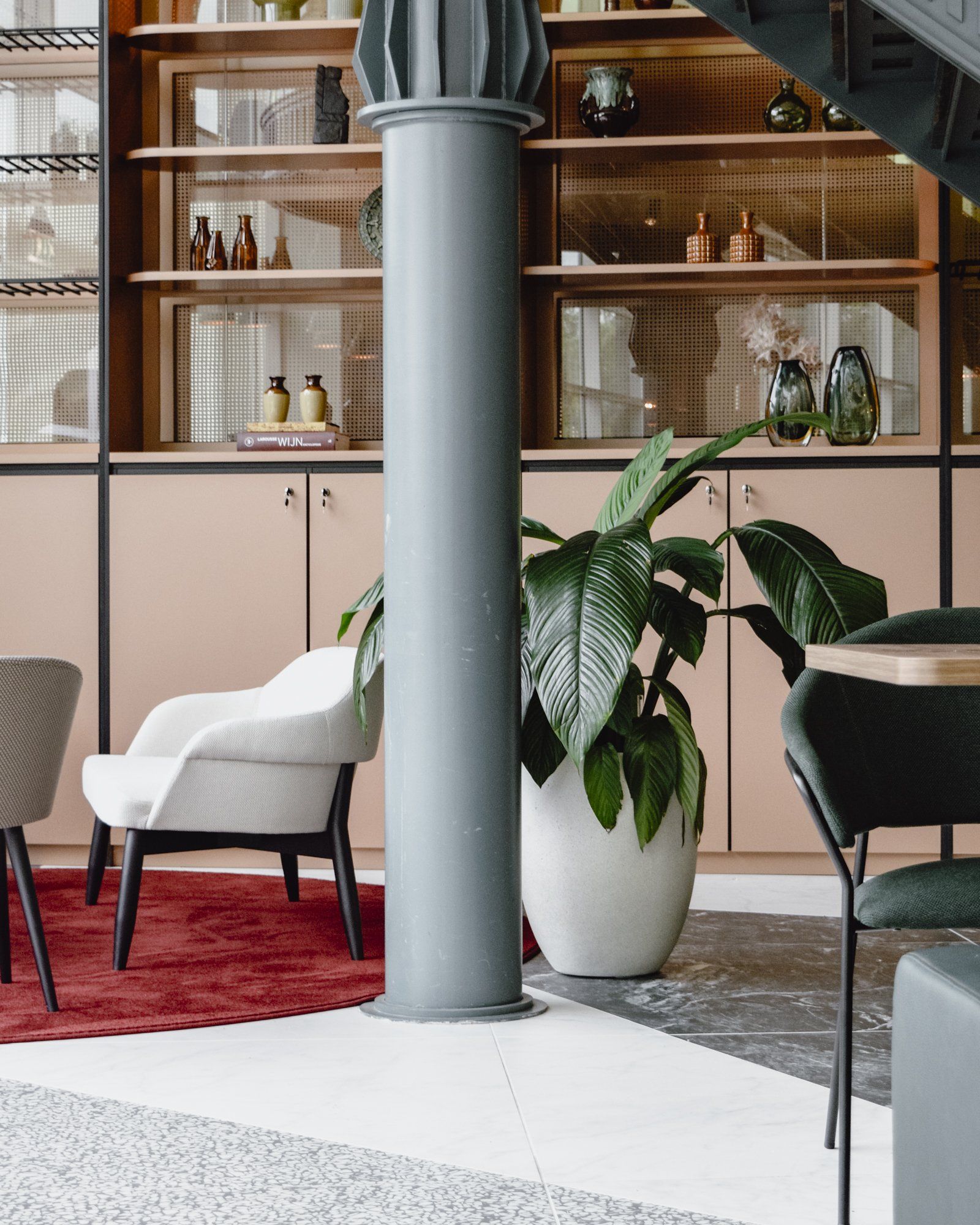 Modern lounge with white chairs, red rug, large potted plant, and wooden shelving around a gray column