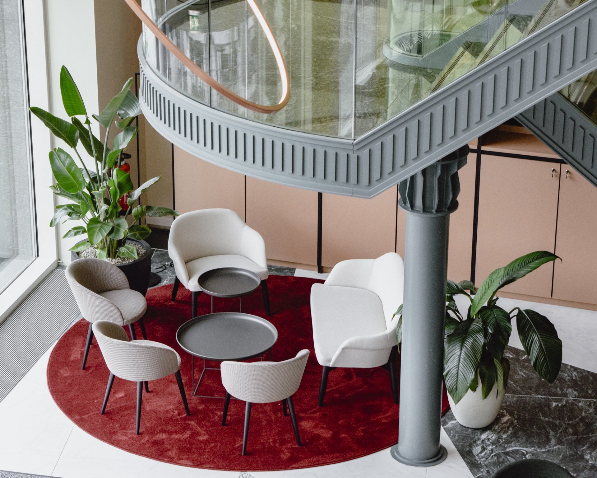 Modern lounge with red rug, white chairs, round table, and indoor plants under a spiral staircase