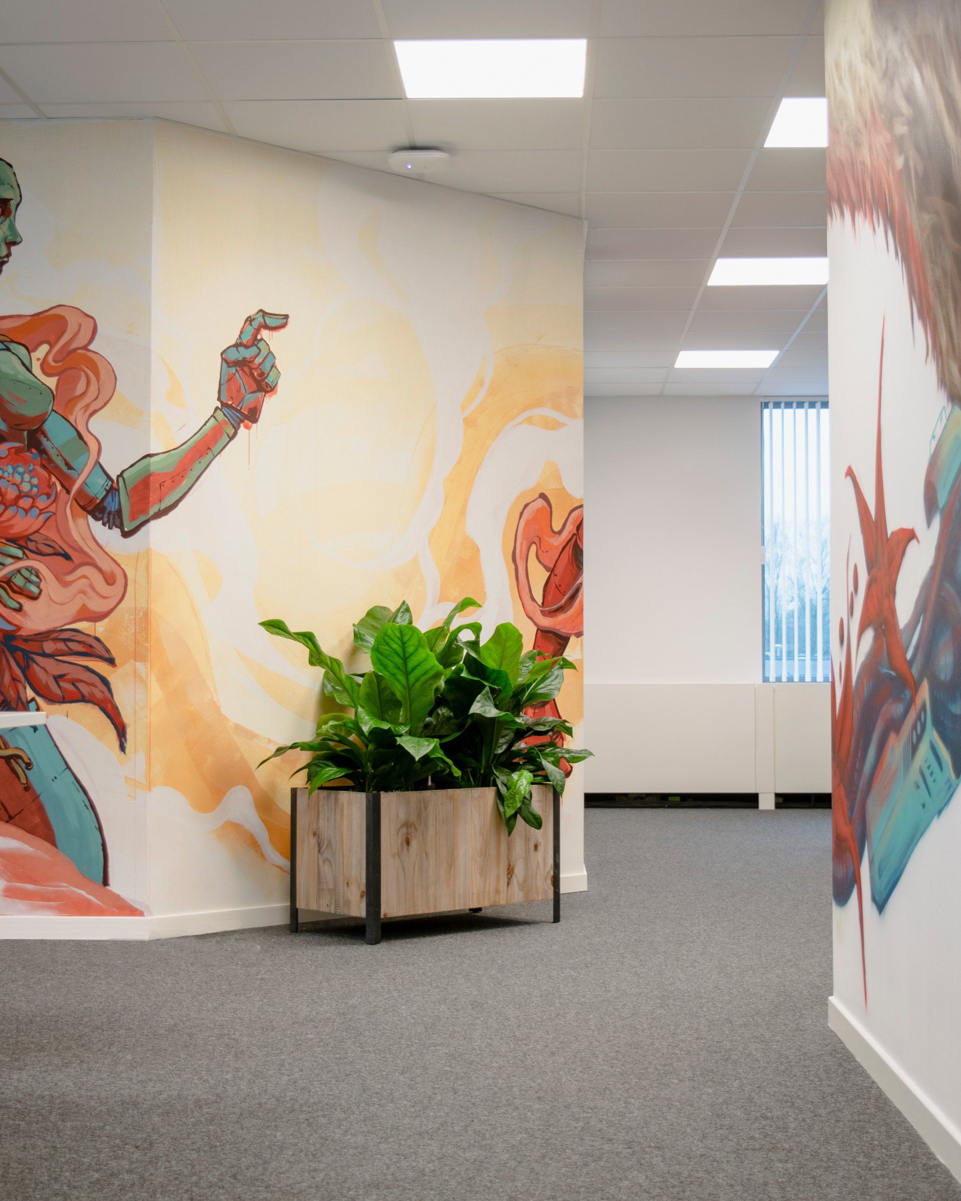 Office hallway with colorful wall murals, gray carpet, and a potted plant in a wooden planter.