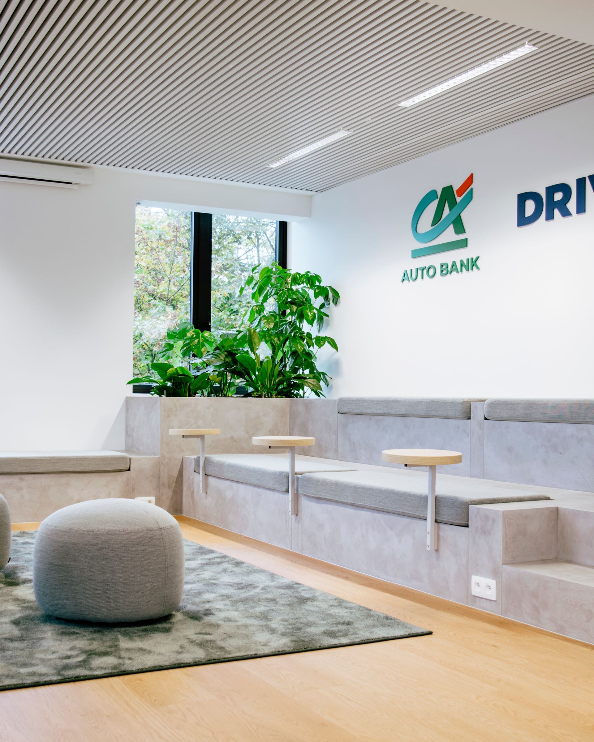 Modern bank lobby with gray seating, green plants, wood flooring, and white walls with logo signage