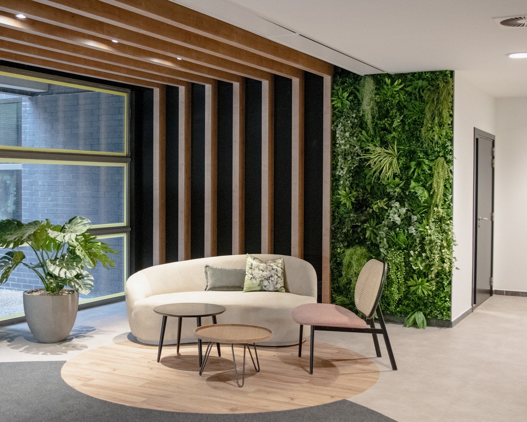 Salon moderne avec canapé incurvé, fauteuils, tapis rond, mur en lattes de bois et mur végétal près de grandes fenêtres