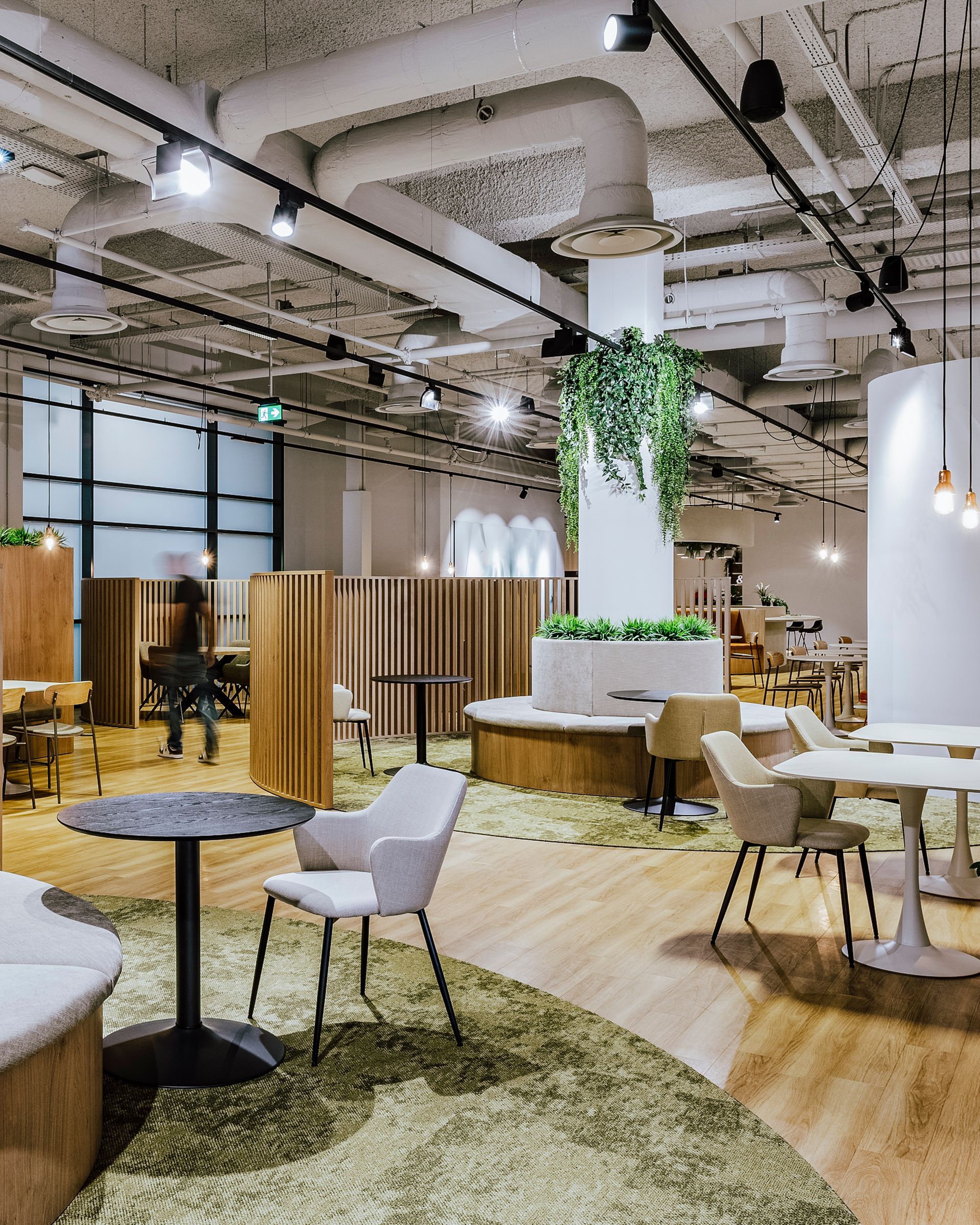 Espace de détente moderne en open space avec sièges en bois, tables rondes, plantes et luminaires suspendus.