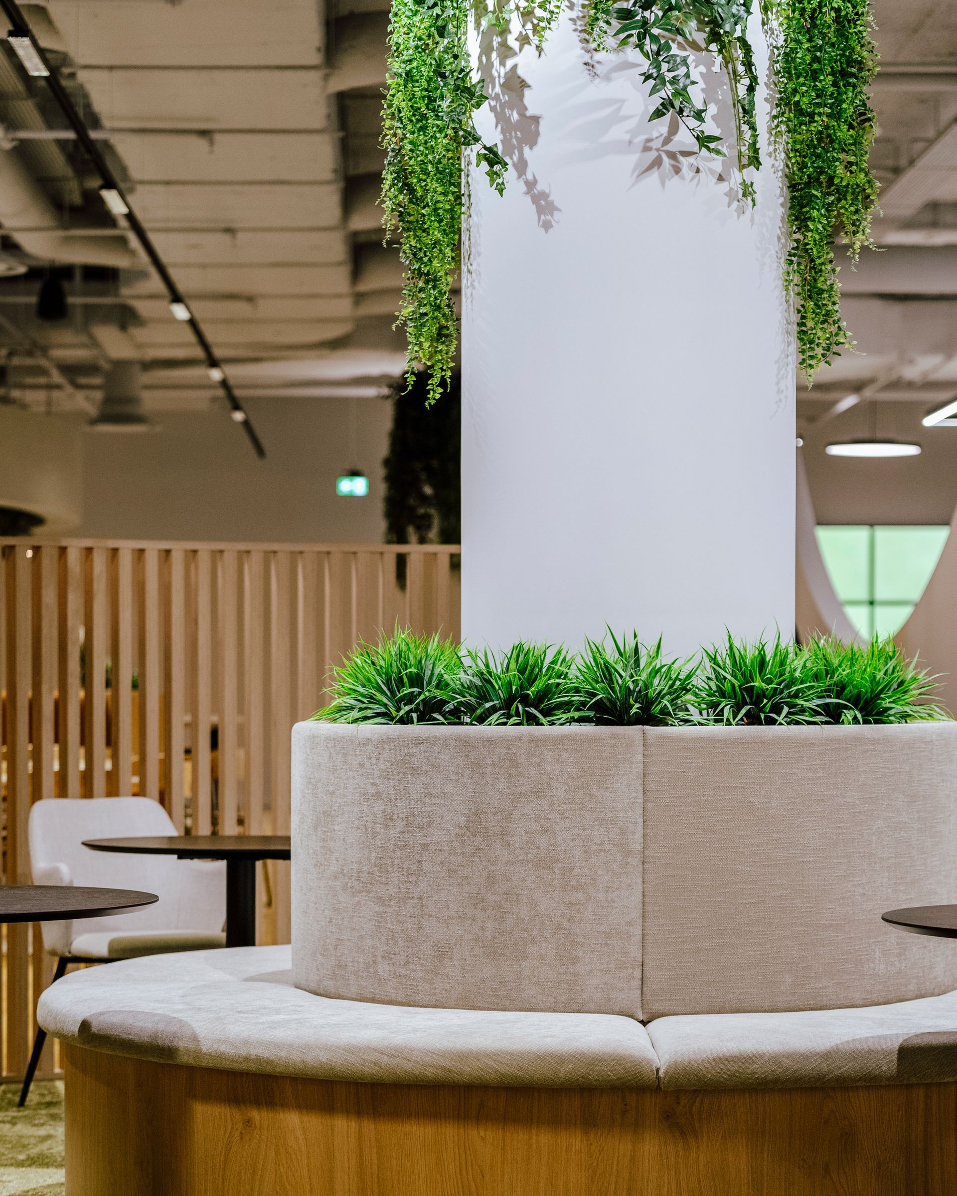 Salon intérieur avec un banc rond beige orné d'une jardinière et de plantes grimpantes vertes enroulées autour d'une colonne blanche.