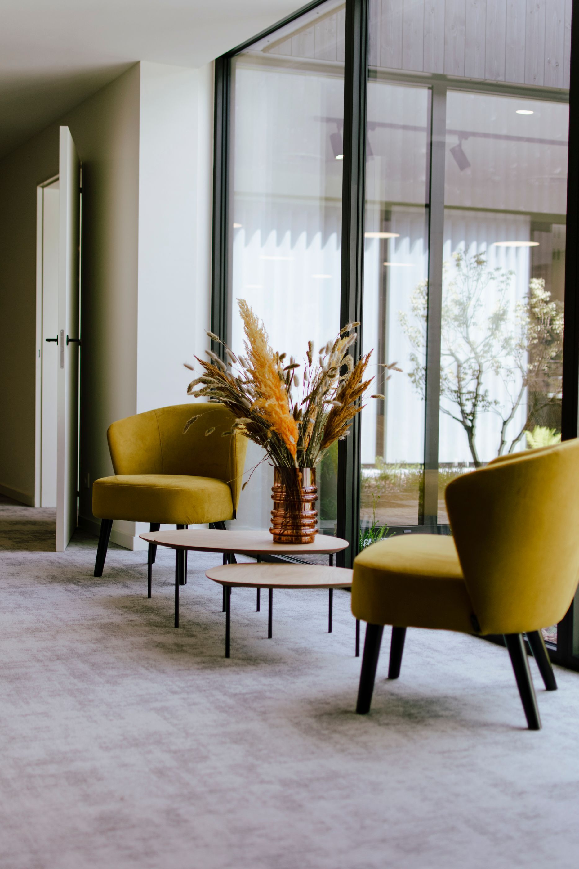 Modern lounge with two olive chairs, low tables, and a vase of dried flowers by large windows