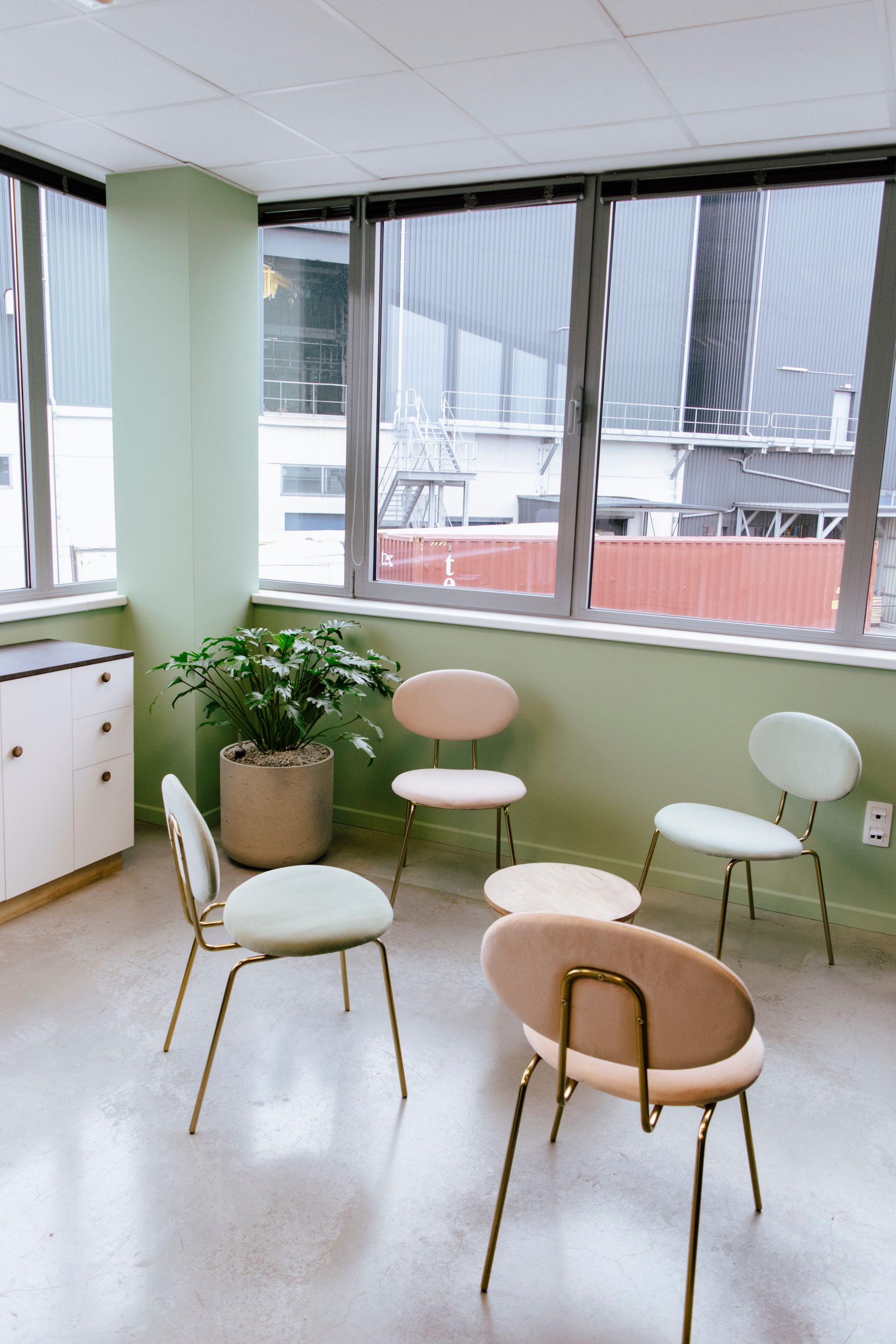 Bright lounge corner with pastel chairs, a potted plant, and large windows overlooking buildings