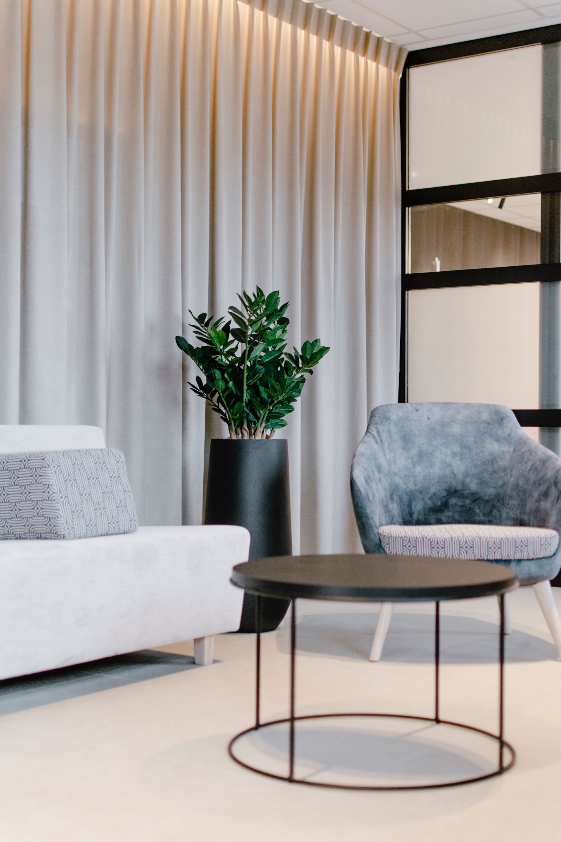 Salon moderne avec canapé blanc, fauteuil gris, table basse ronde et plante en pot près des rideaux.