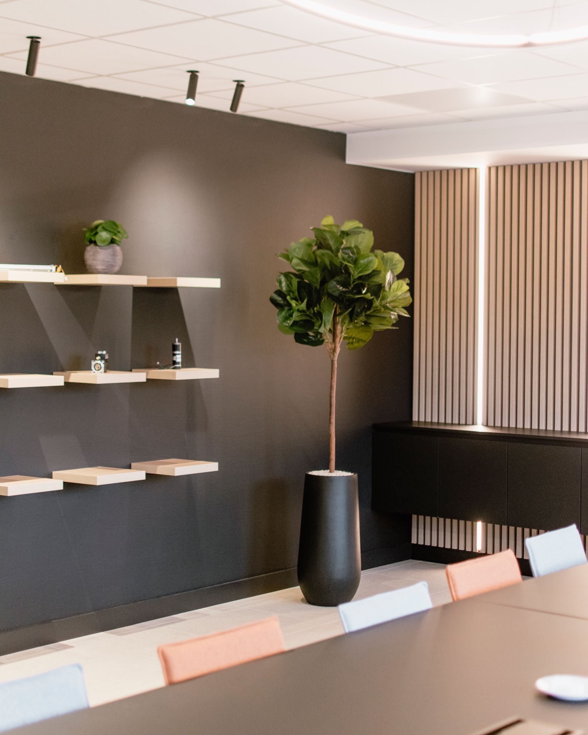 Salon de bureau moderne avec étagères murales, arbre en pot et stores rayés derrière une table de conférence