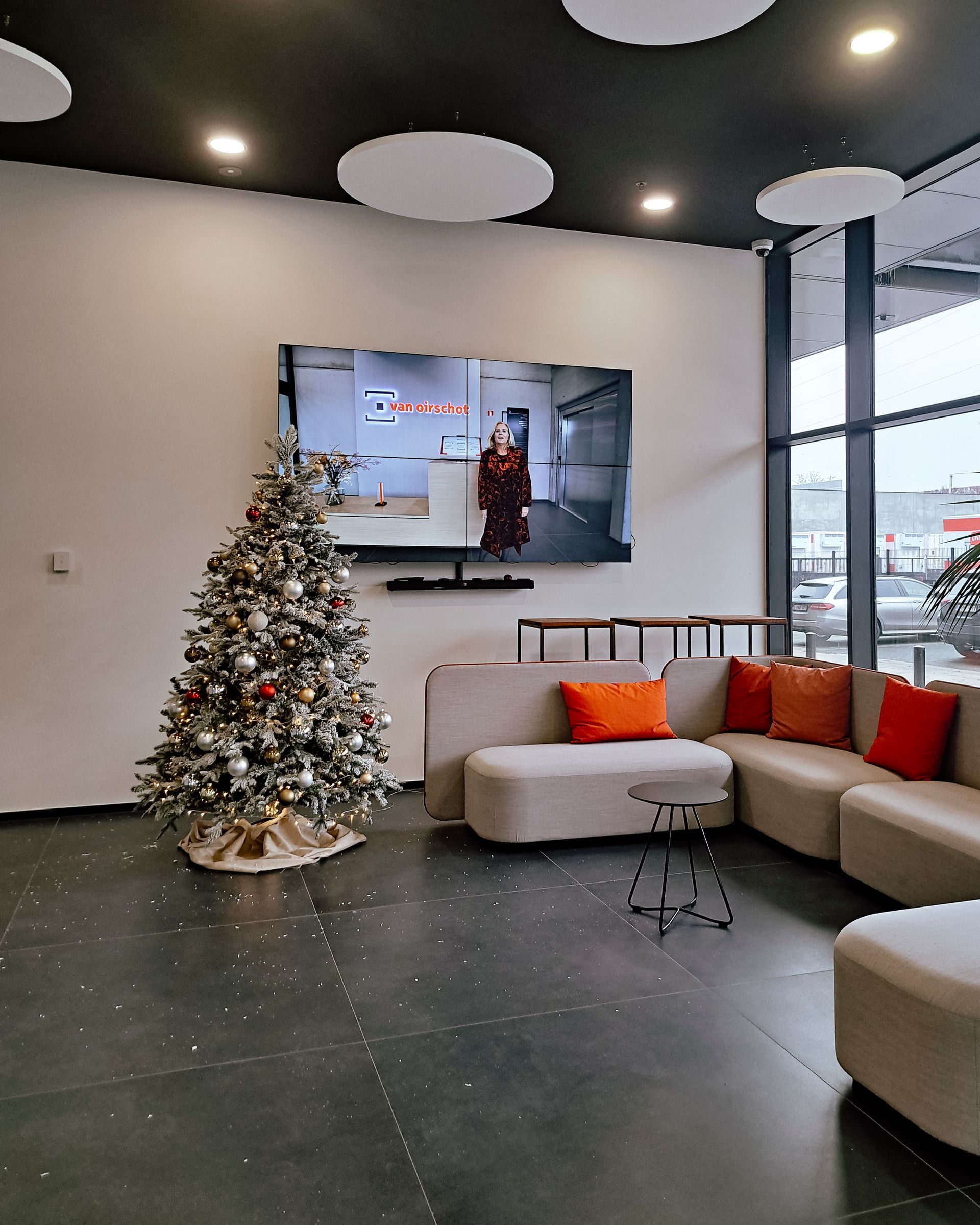Modern lobby with a decorated Christmas tree, gray sectional sofa, wall TV, and large windows.