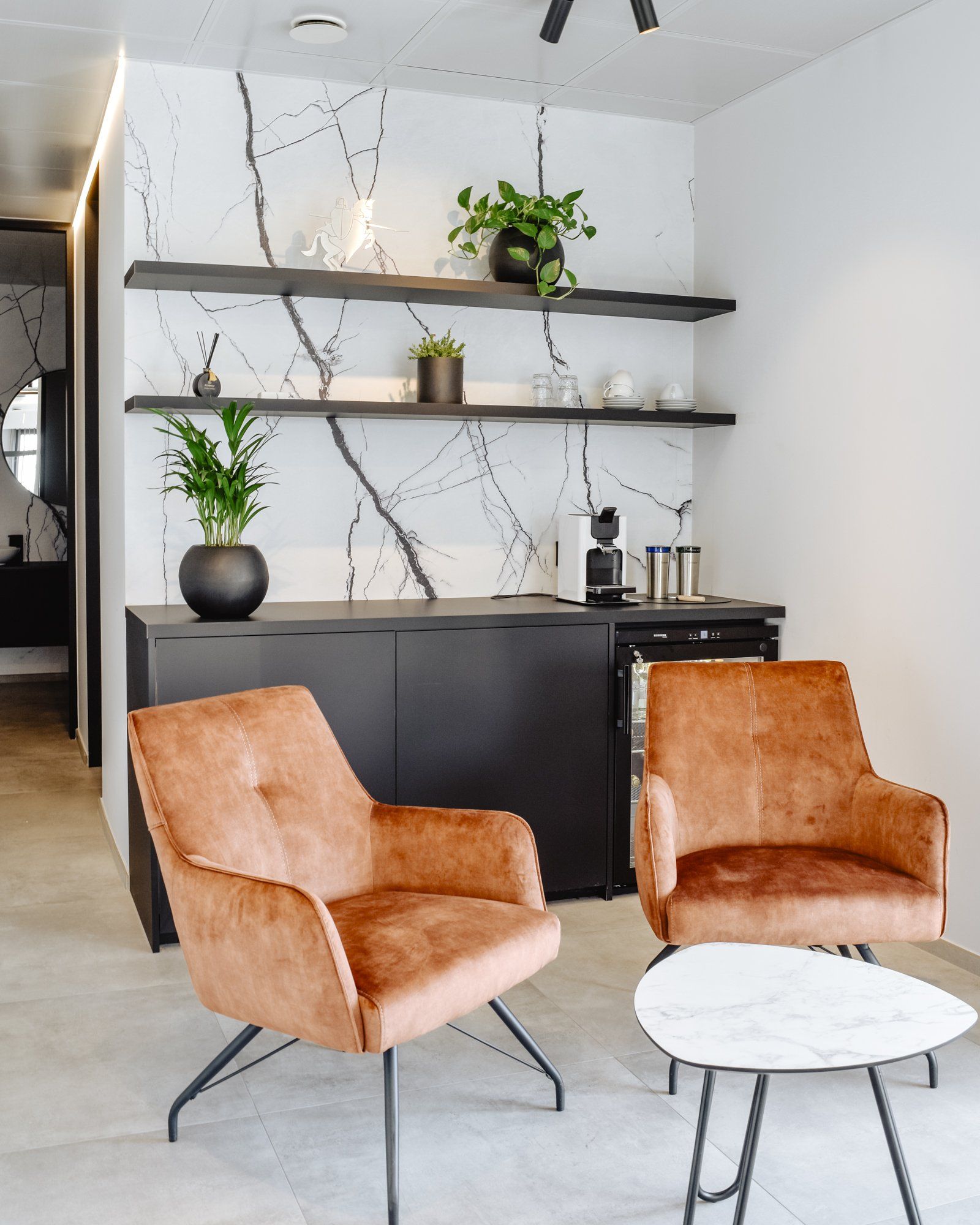 Modern living room with two tan chairs, round white table, black cabinet, and wall shelves with plants.