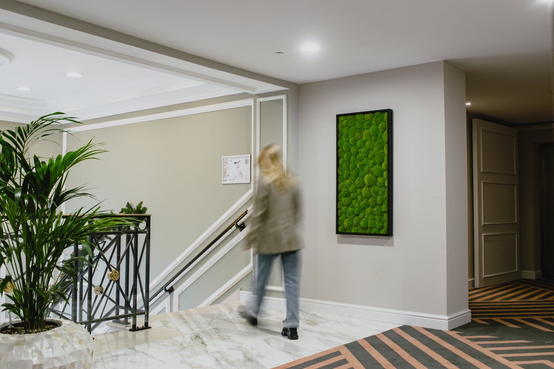 Personne floue marchant dans un couloir moderne, à côté d'un escalier et d'une œuvre d'art murale verte.
