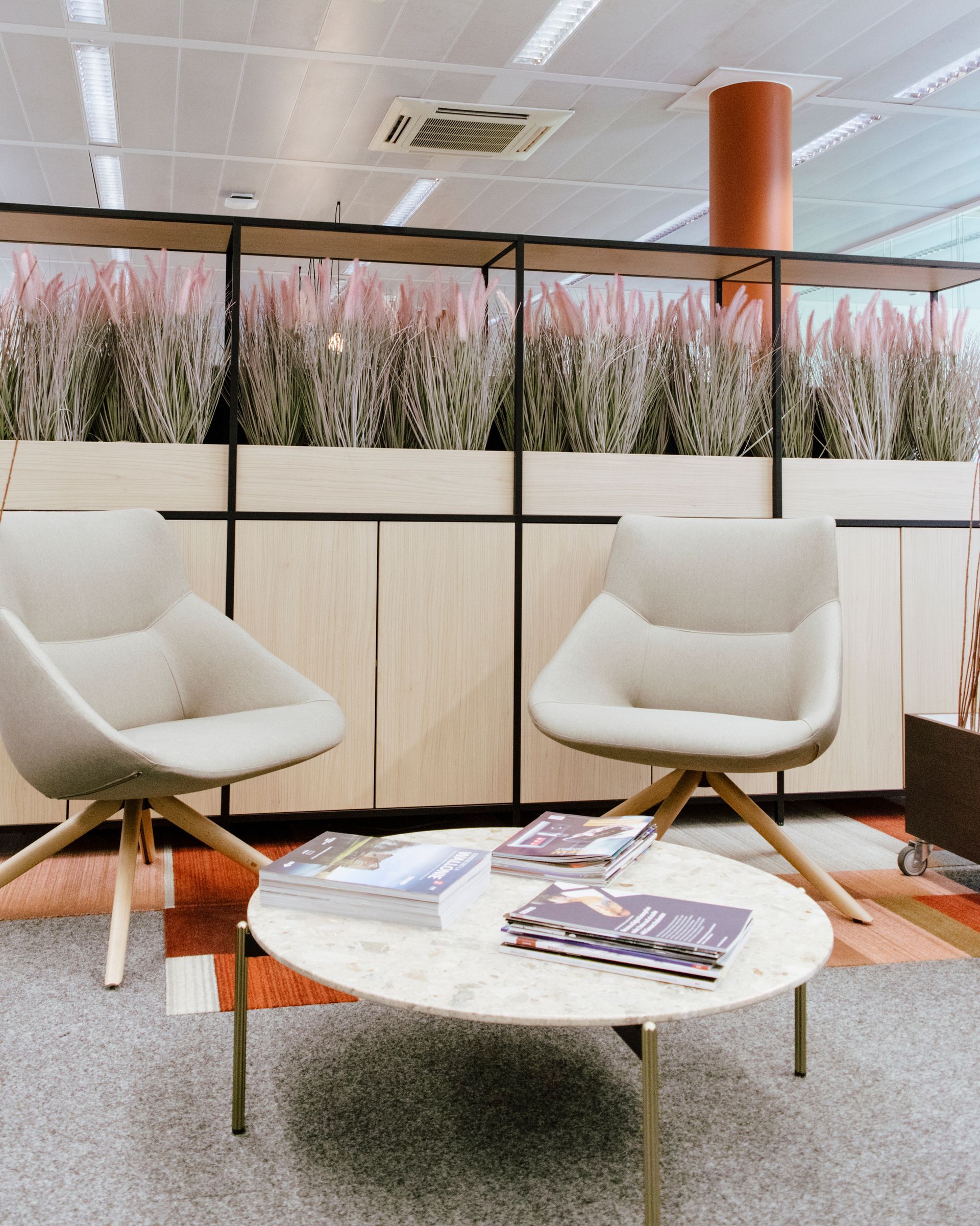 Modern lounge with two white chairs, round coffee table, and tall planter partition under a bright ceiling