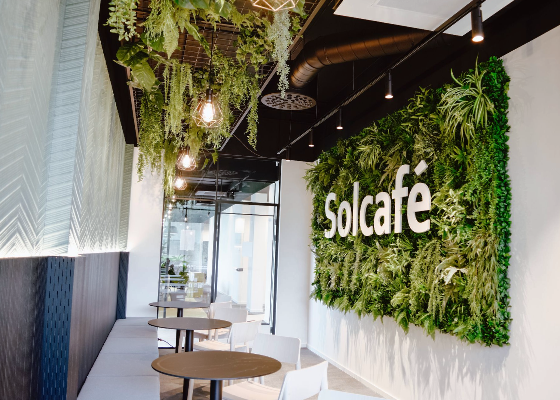 Het interieur van een cafetaria bij Soltech, met een weelderige verticale plantenwand, witte tafels en hangende planten.