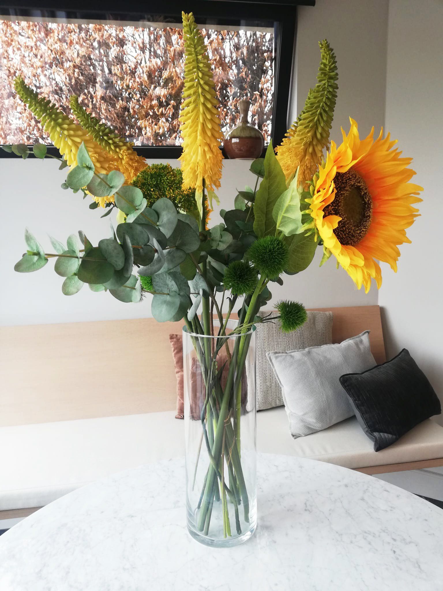 Sunflowers and eucalyptus in a glass vase on a white table in a bright living room.