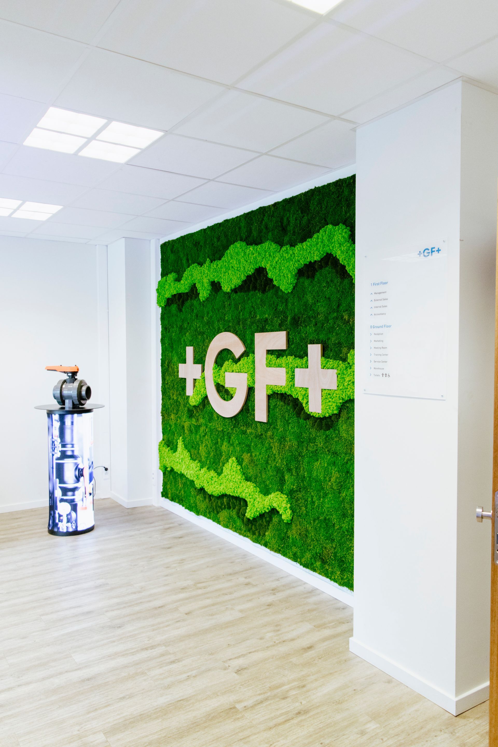 Couloir de bureau avec un mur recouvert de mousse verte affichant « +GF+ » et une sculpture bleue décorative à proximité