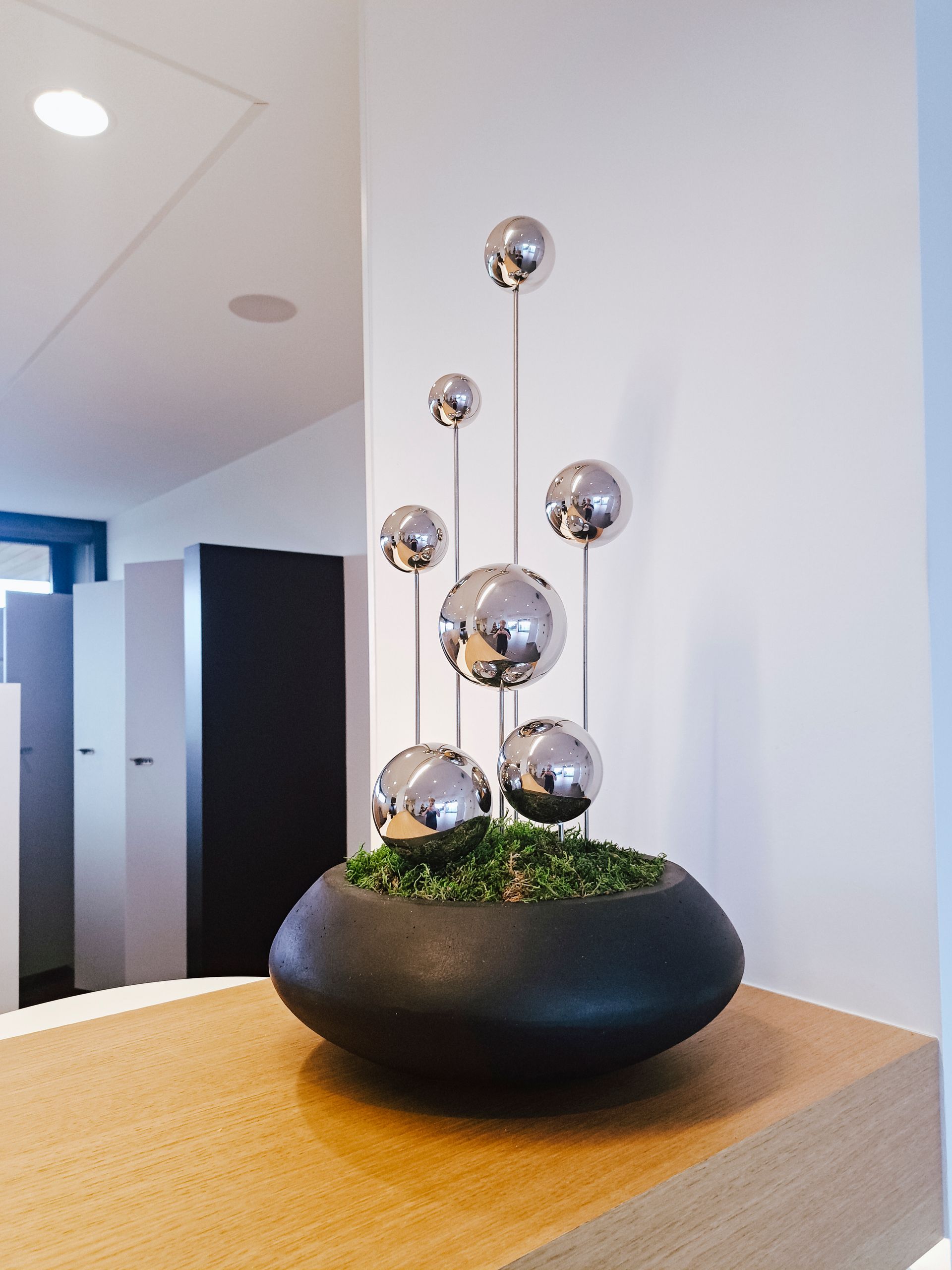 Modern metal sculpture with chrome spheres in a black bowl planter on a wooden table in an office hallway