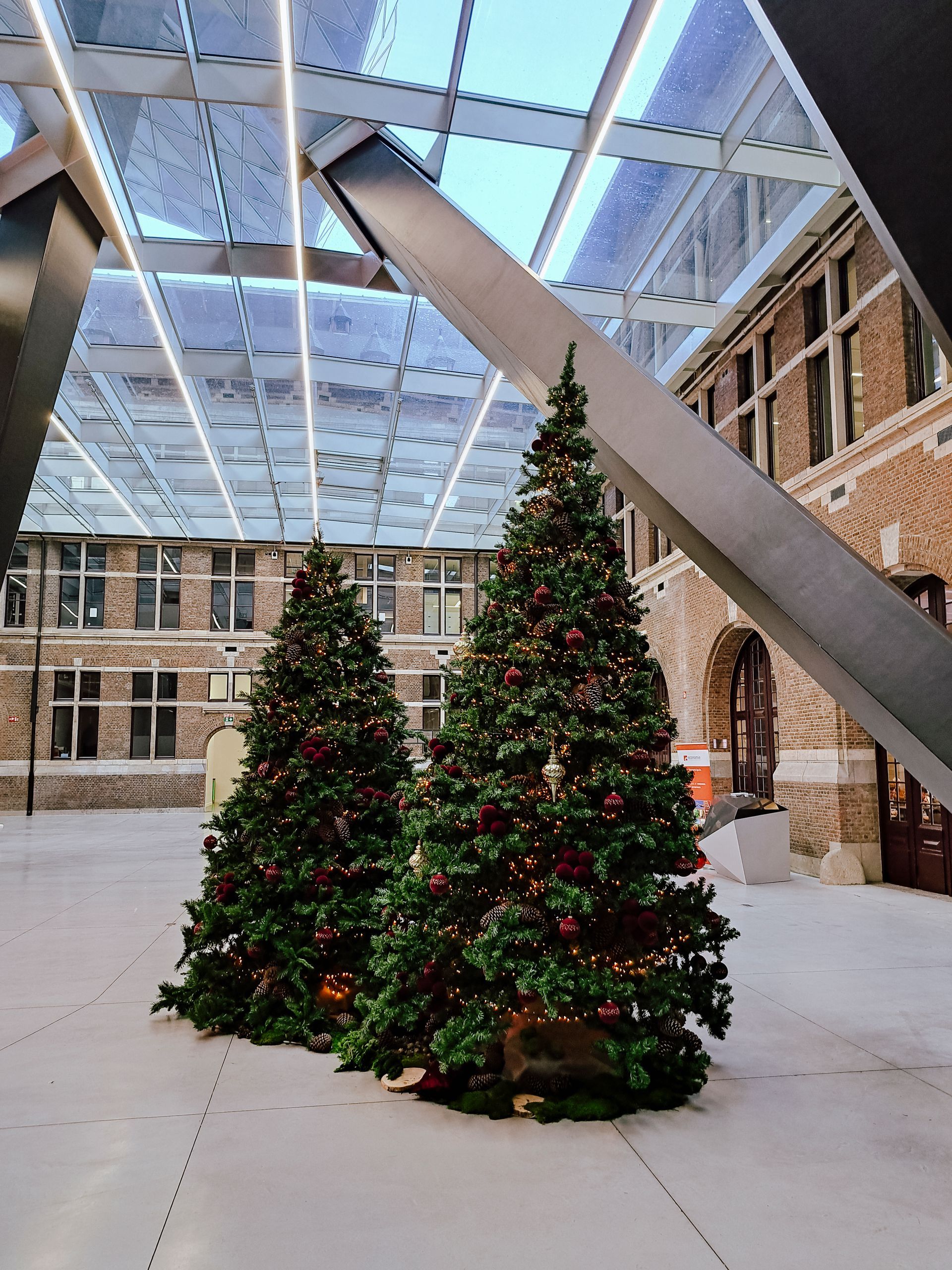 Two lit Christmas trees in a covered atrium with brick walls and a glass roof