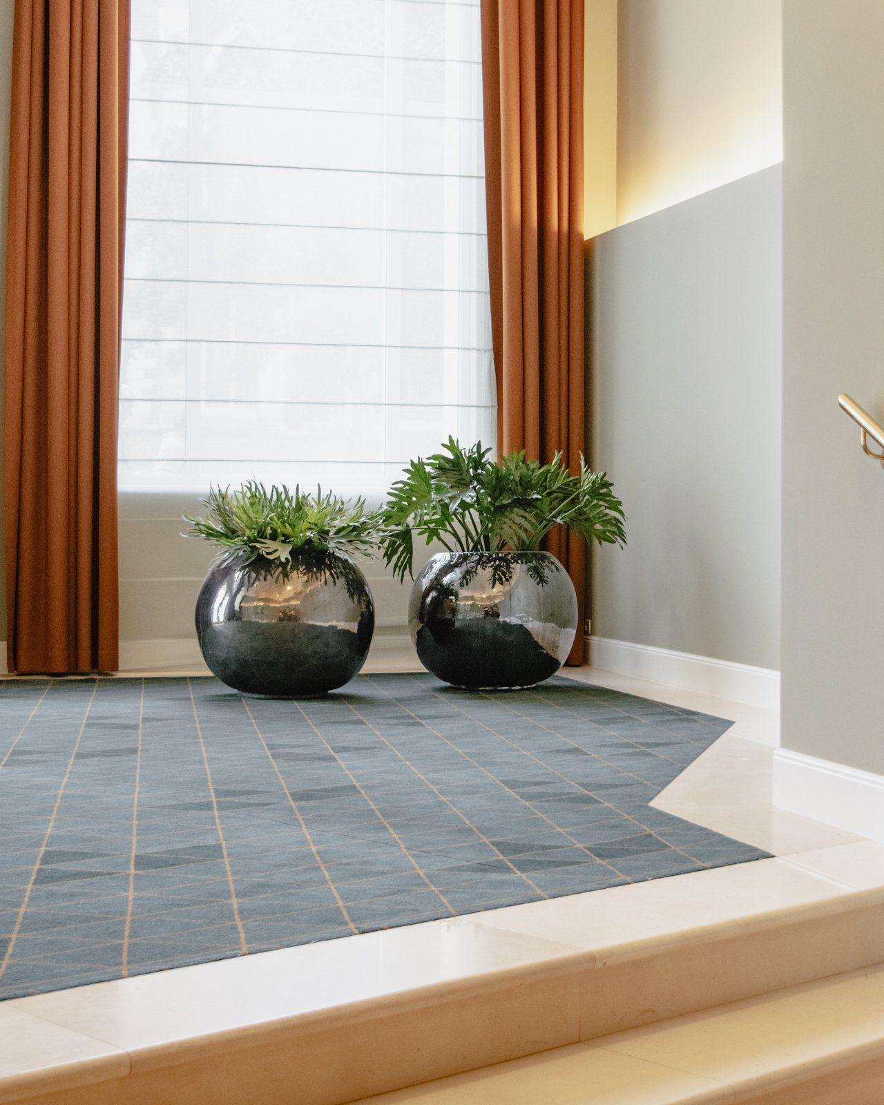 Two potted plants on a blue rug in a bright entryway with orange curtains and a white wall