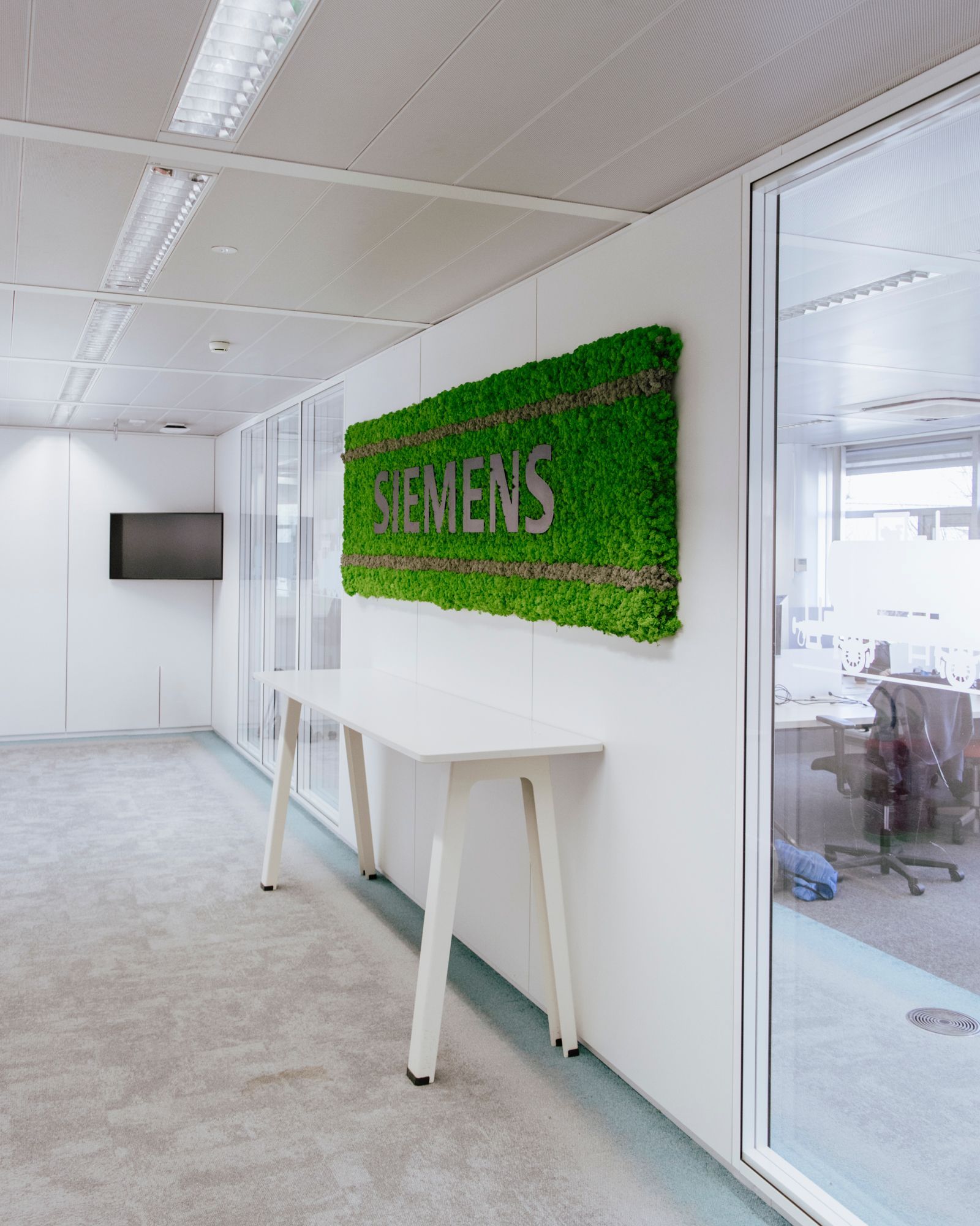 Couloir de bureau lumineux avec une enseigne murale verte Siemens et une table blanche à côté de portes vitrées.