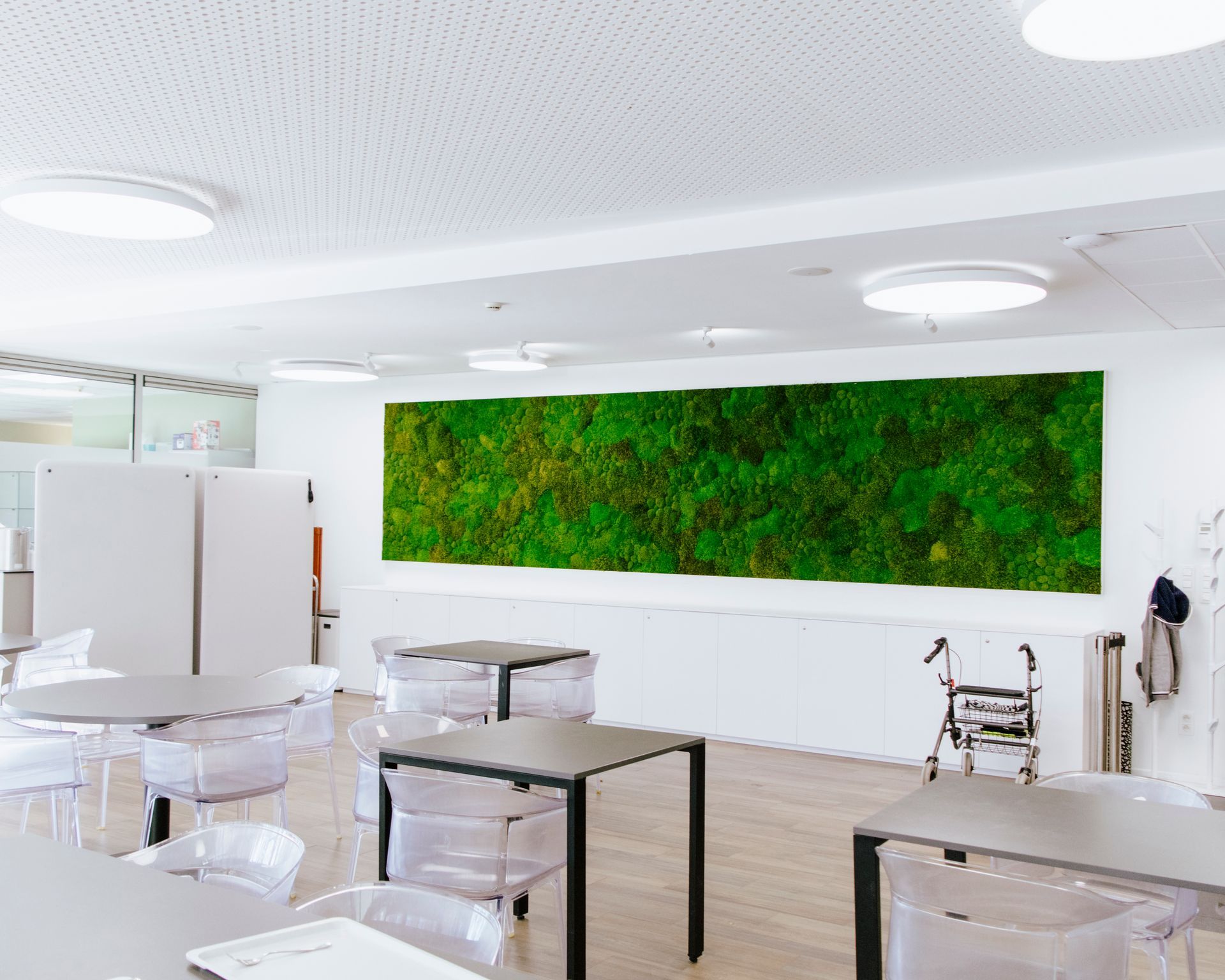 Salle de classe lumineuse avec des tables et des chaises blanches, et un grand mur végétal vert au fond.