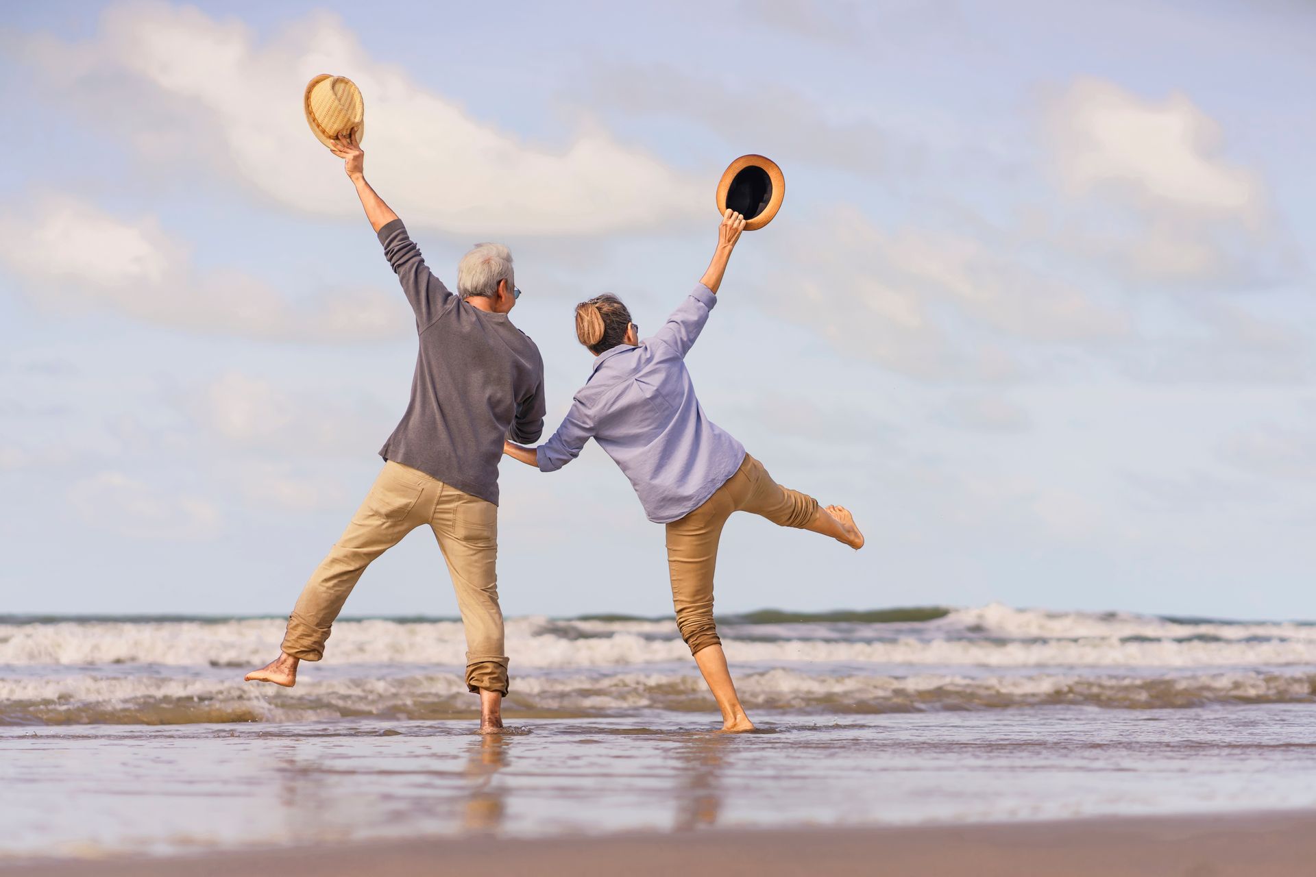 Two people on a beach jump with arms raised, holding paddles, under a cloudy sky.