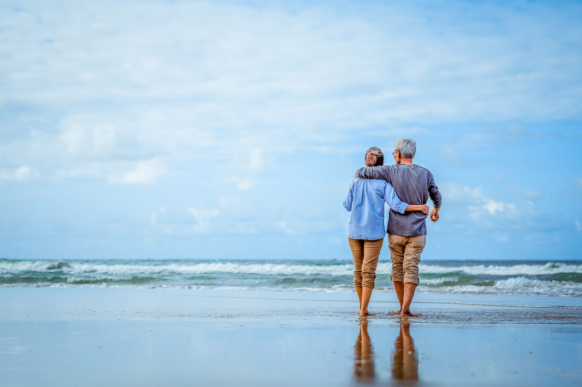 Two people stand arm in arm on a beach, facing the ocean under a blue sky.
